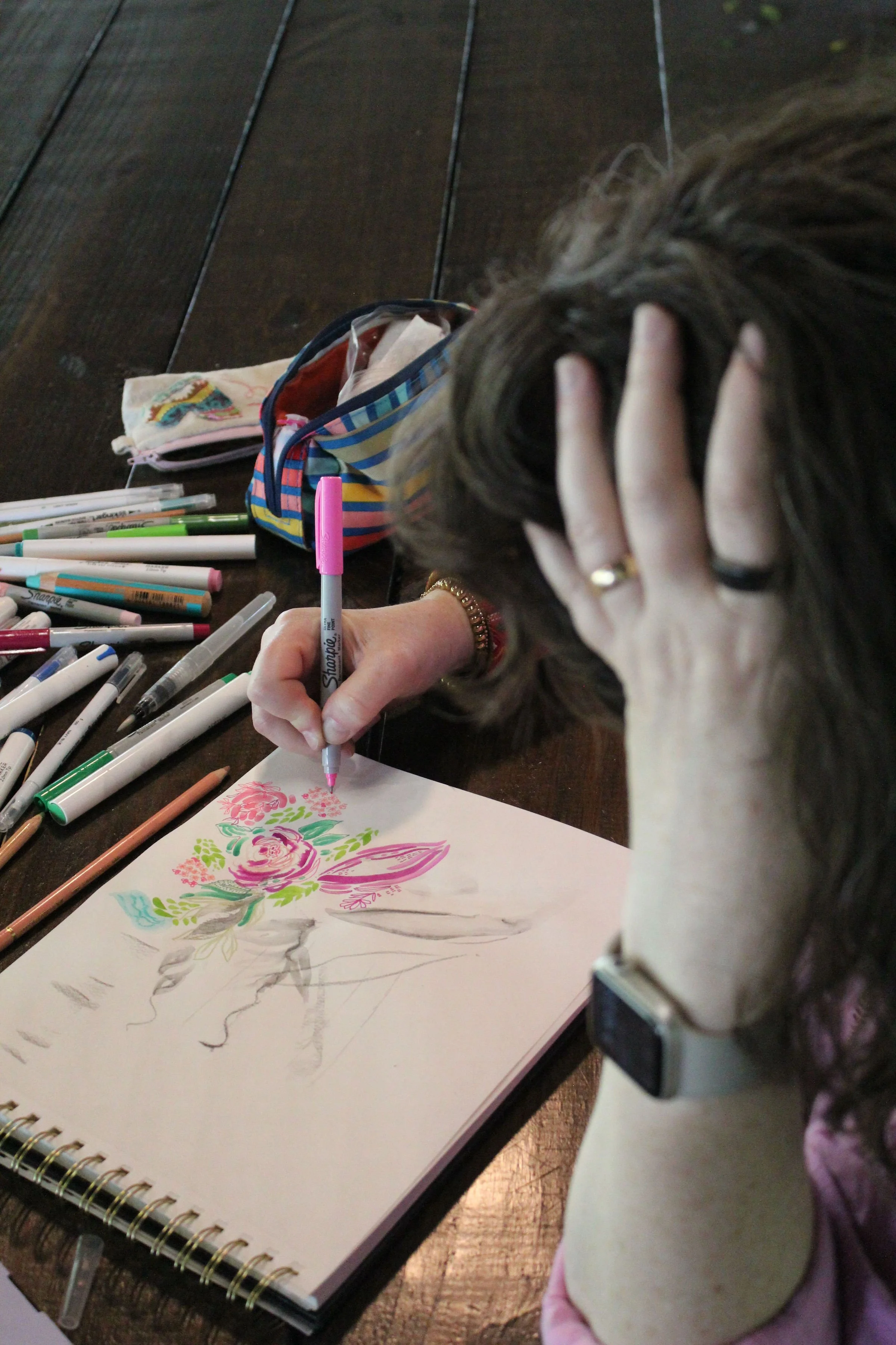 A person with brown hair, wearing a ring on their finger, is drawing on a sketchbook with colored markers. The sketchbook has colorful floral drawings. There are numerous markers scattered on a dark wooden surface. The person's hand rests on their head, and they are wearing a white watch.