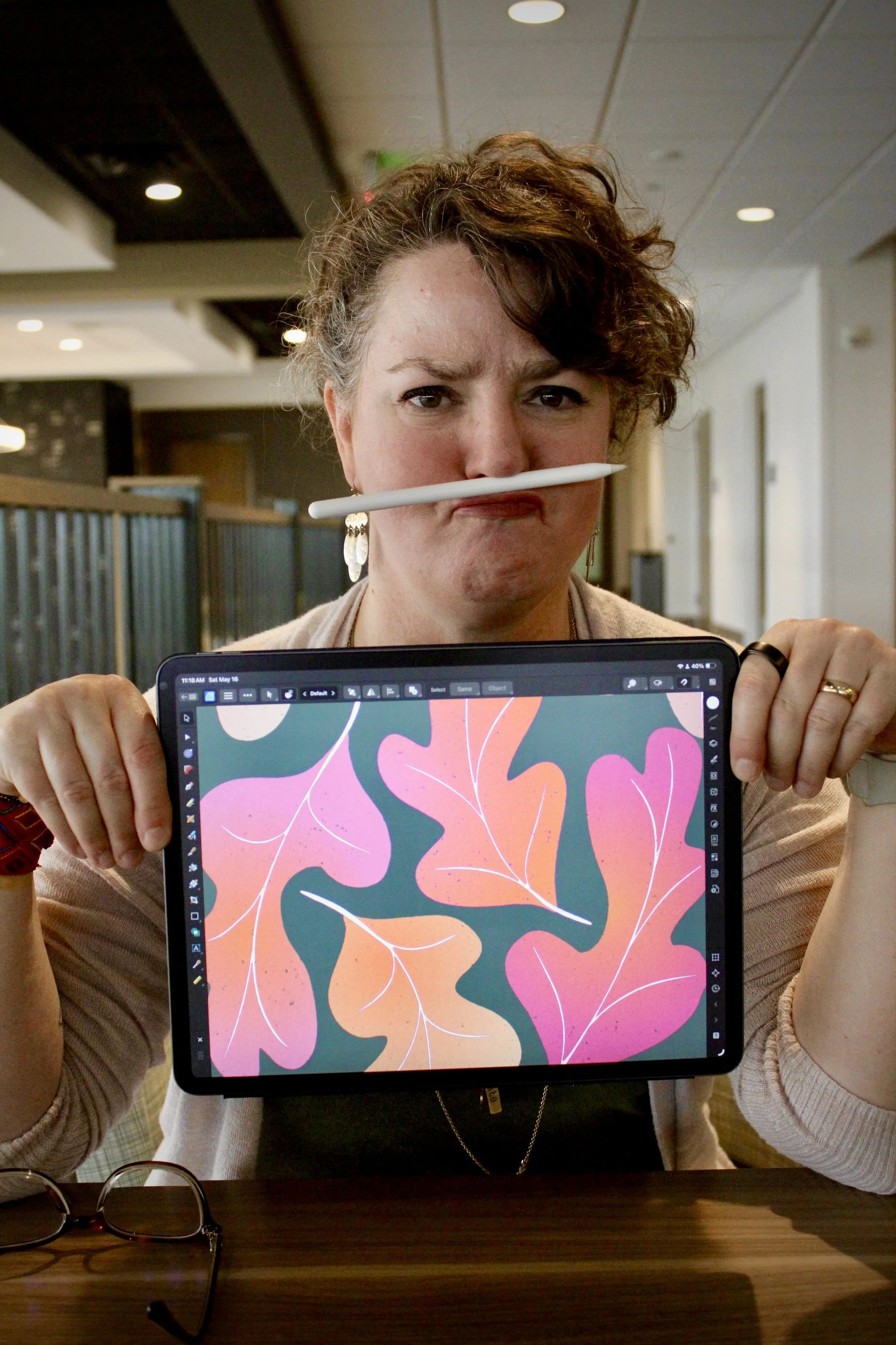 A woman with curly hair up in a ponytail, wearing earrings, is holding a tablet with a colorful abstract design of fall leaves on the screen. She is indoors, sitting at a table, with glasses placed on the table in front of her.