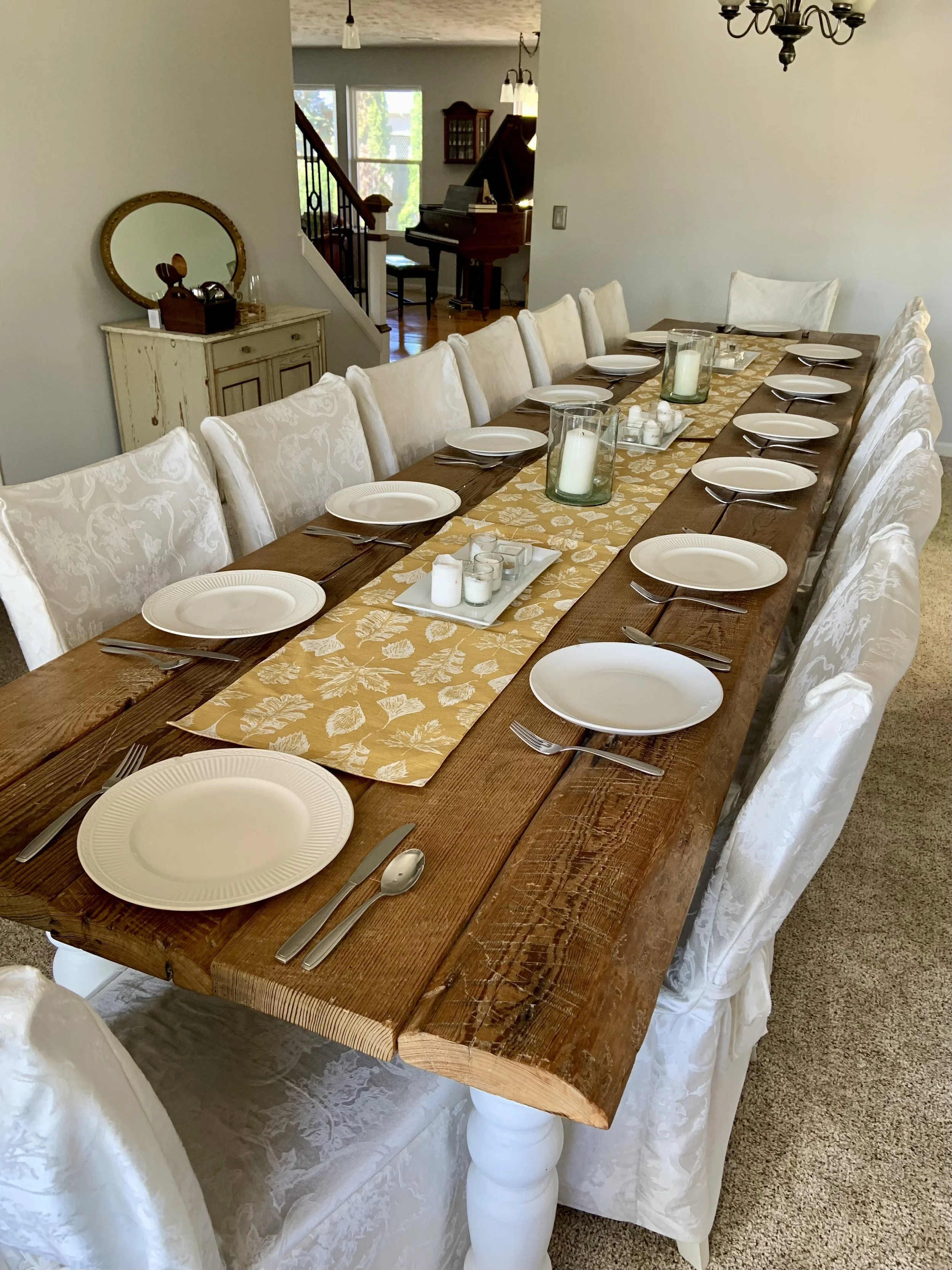 A long wooden dining table set for a meal with white plates, silverware, and candles in glass holders. The table has a yellow and white floral table runner, and white upholstered chairs with slipcovers surround it. In the background, a sideboard with decorative items and a mirror is visible, along with a staircase, a piano, and a window with natural light.