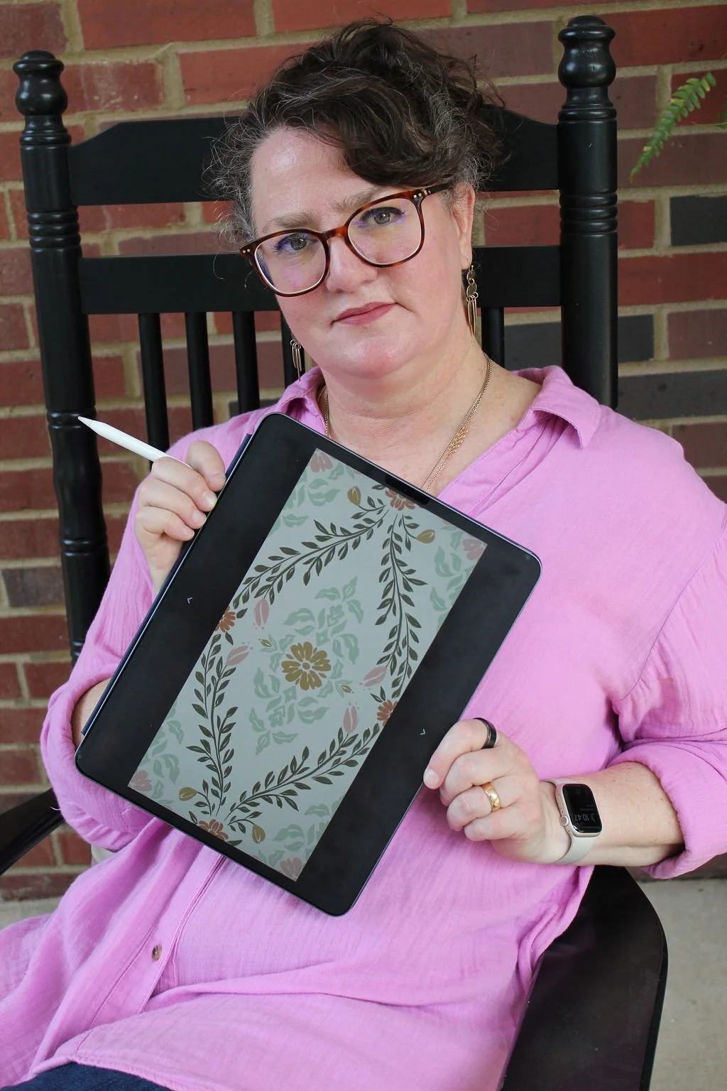 A woman wearing glasses and a pink shirt sitting on a black chair in front of a brick wall, holding a digital tablet with a floral design on the screen and a stylus in her right hand.