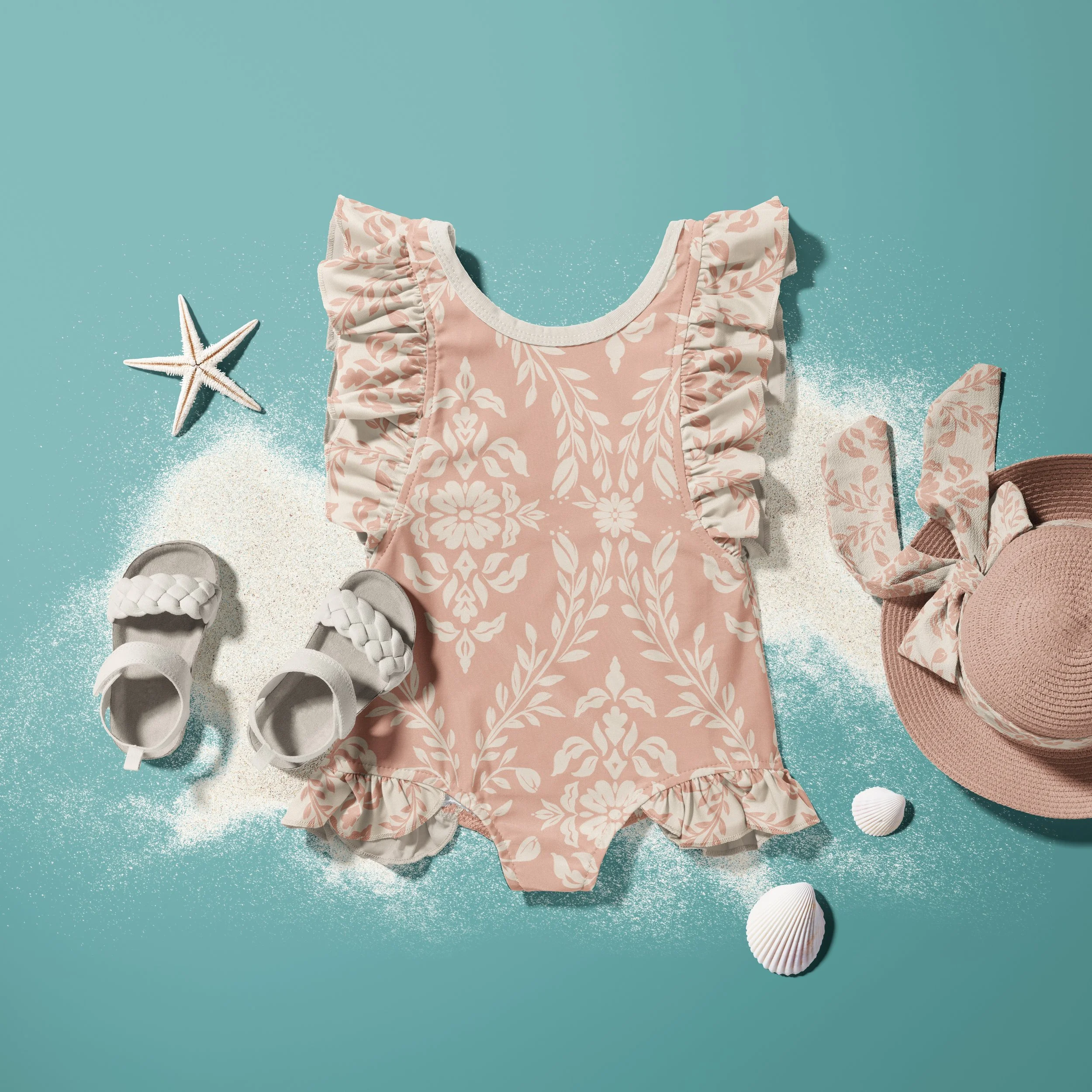 A baby onesie in a pink and white patterned fabric with ruffled sleeves, starfish, seashells, and sand on a blue background, suggesting a beach or seaside theme.