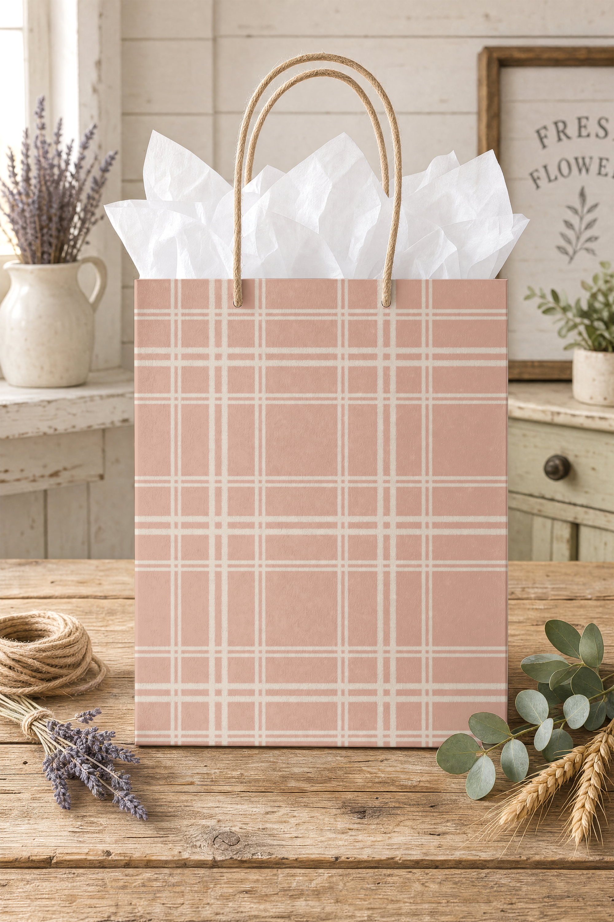A pink gift bag with a white plaid pattern, filled with white tissue paper, placed on a rustic wooden table with lavender, eucalyptus, and wheat beside it. A white wooden wall and framed floral print are in the background.