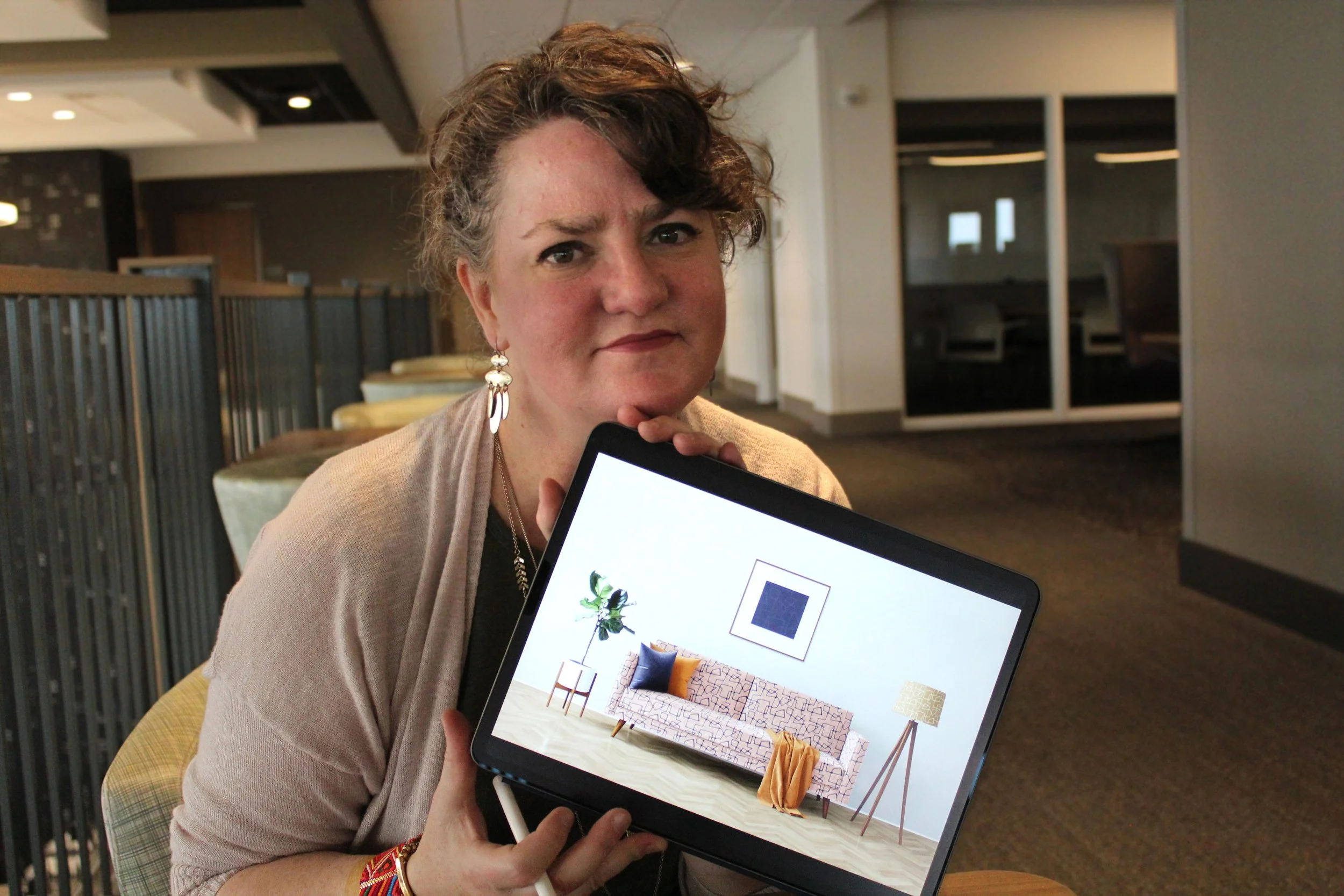 A woman with short curly hair and earrings holding a tablet with interior design images inside.