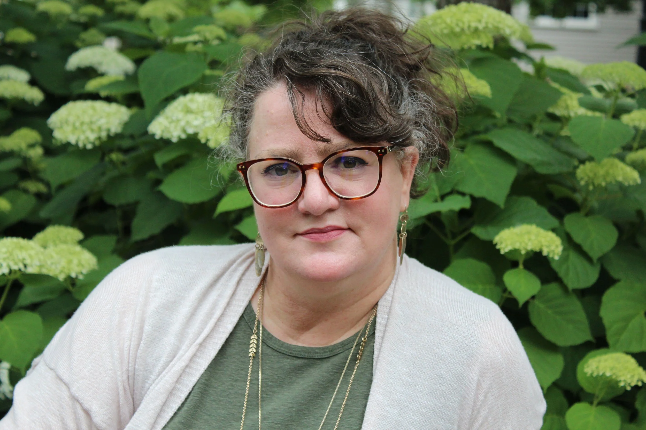 A woman with short curly hair and glasses, wearing earrings, a necklace, and a beige cardigan over a green shirt, standing outdoors in front of green hydrangea flowers and leaves.