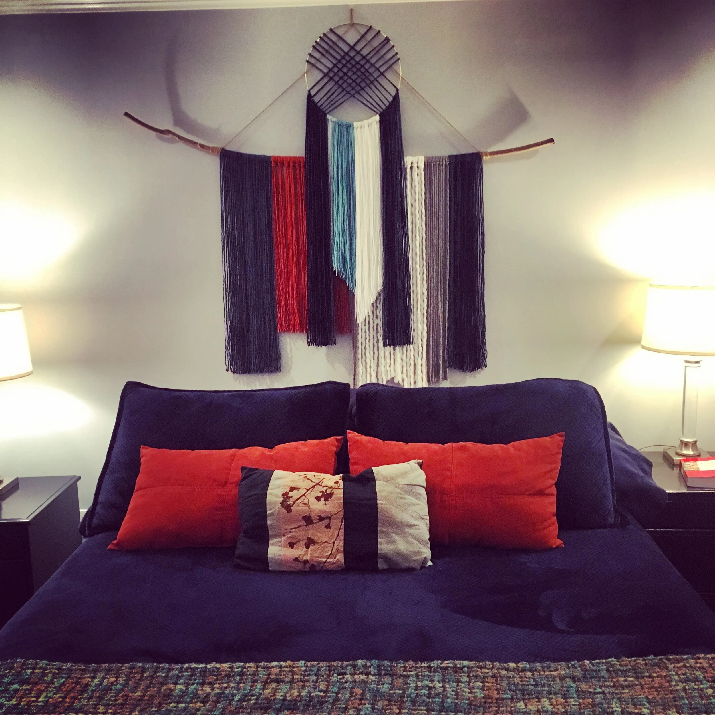 A colorful wall hanging art piece made of vertical strands of yarn in orange, navy, white, gray, and turquoise, attached to a curved branch on a plain white wall, over a navy blue bed with two table lamps on side tables visible below.