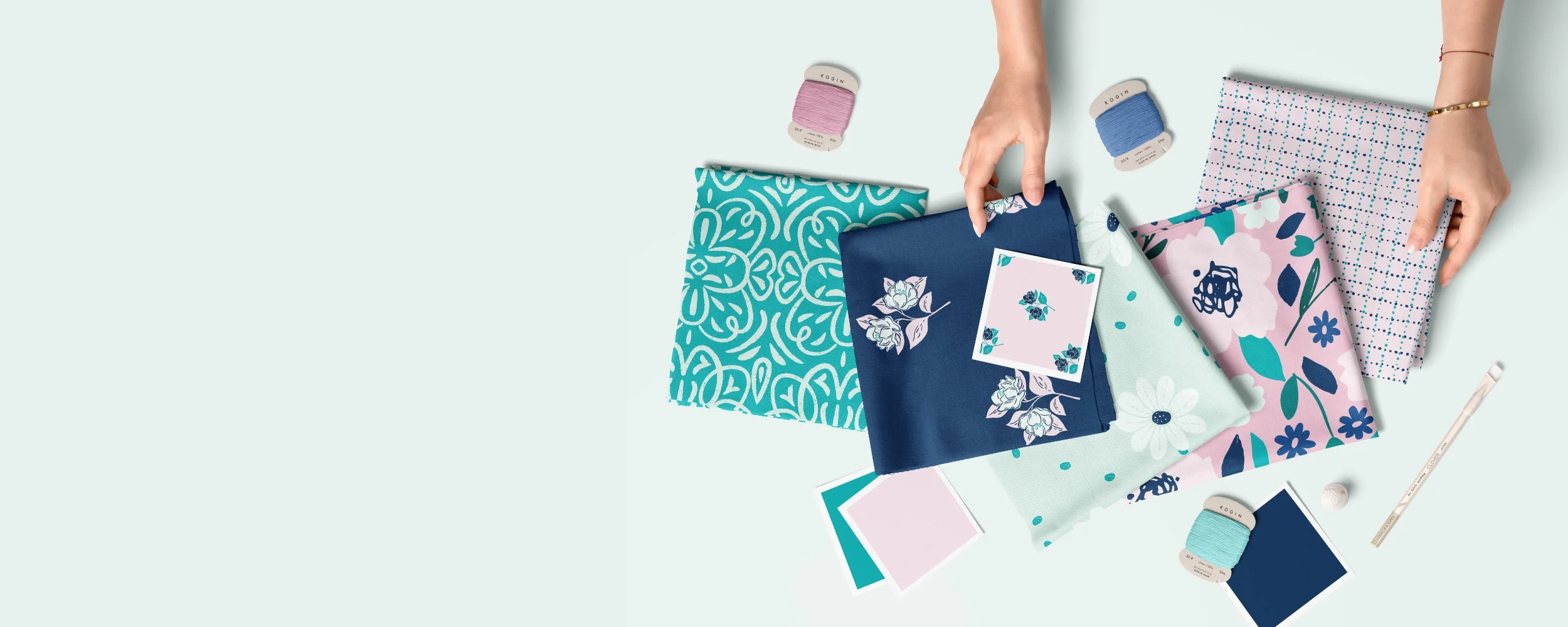 Flat lay of various fabric pieces, threads, and sewing tools in pastel and floral designs, with hands arranging fabric in shades of navy, turquoise, mint, and light pink.