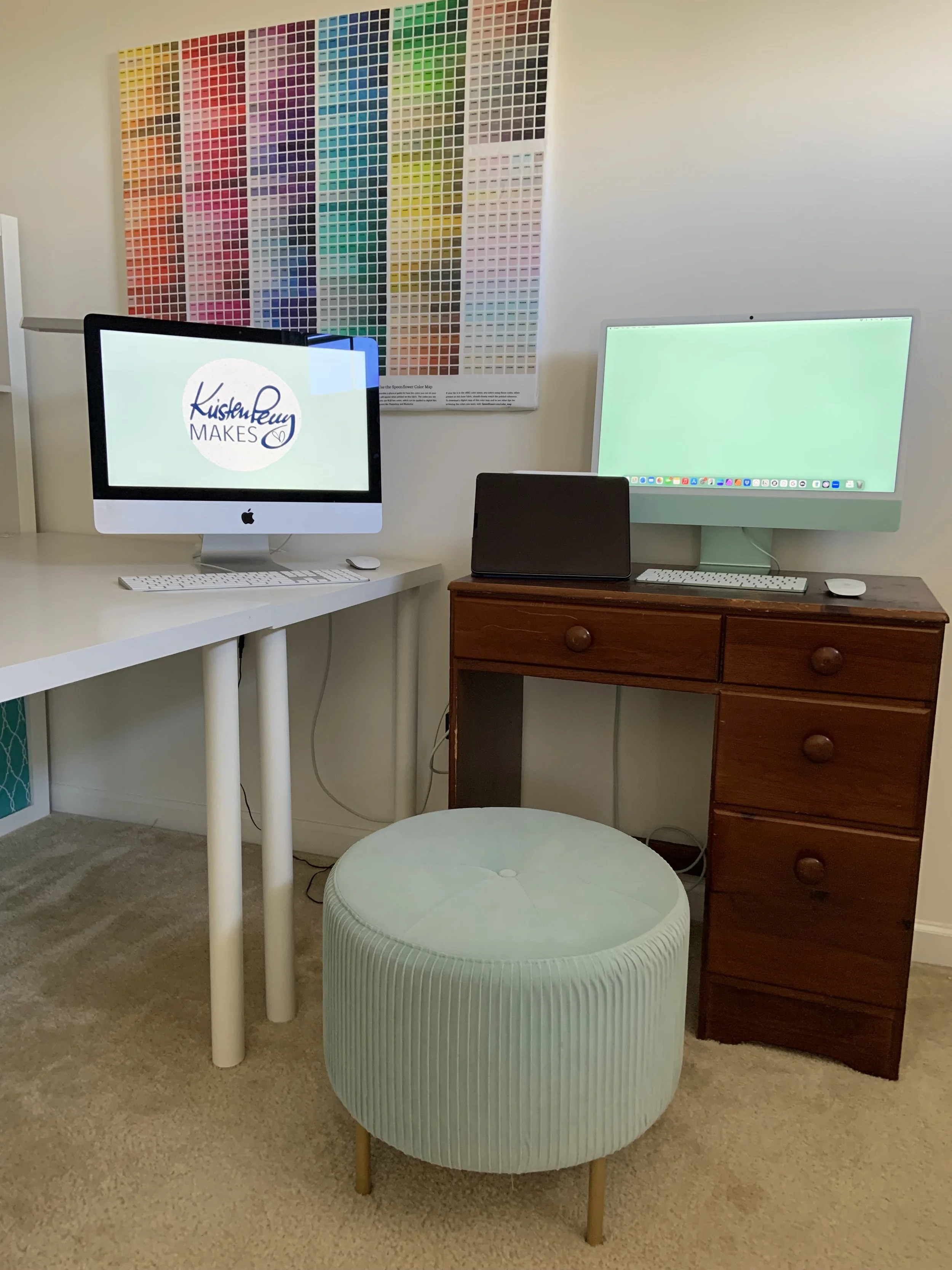 A room with a large color palette poster on the wall, two desktop computers (one with a white frame and the other with a light-colored frame), a small black laptop, a white keyboard, and a white mouse. There is a round, light green upholstered ottoman in front of the computers and dark wooden furniture supporting the devices.