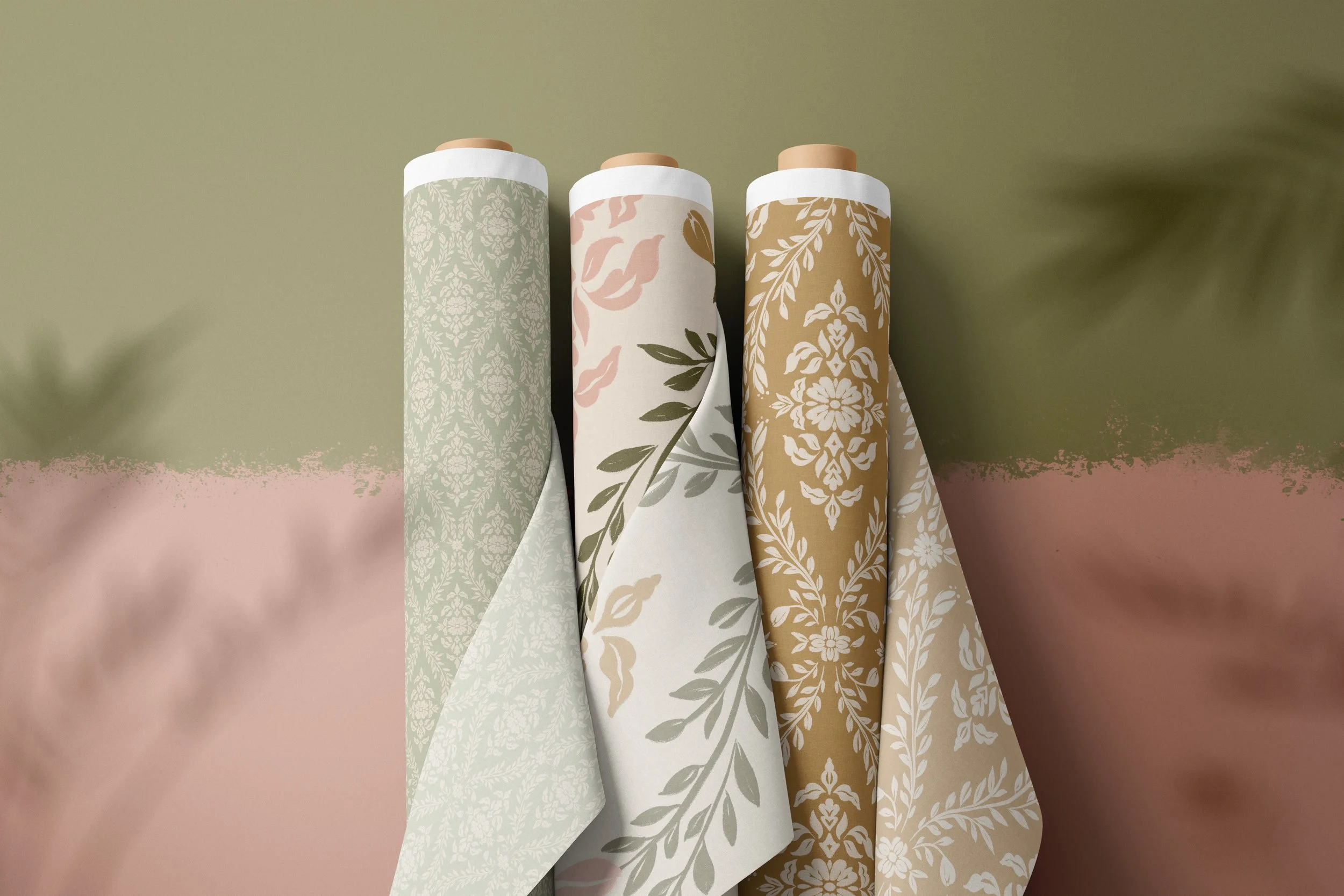 A mockup of three fabric rolls leaning against a pink and green wall.  The fabrics are damask floral patterns in light sage, olive and pink on off white, and sand on deep yellow.