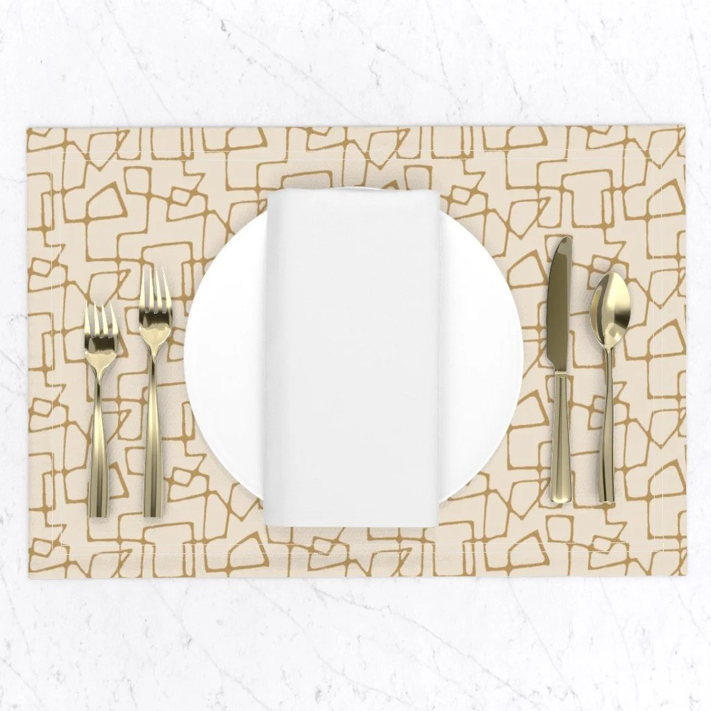 Table setting with gold utensils, a white plate, a folded white napkin, on a cream placemat with an ochre mid-century line pattern, set on a white surface.