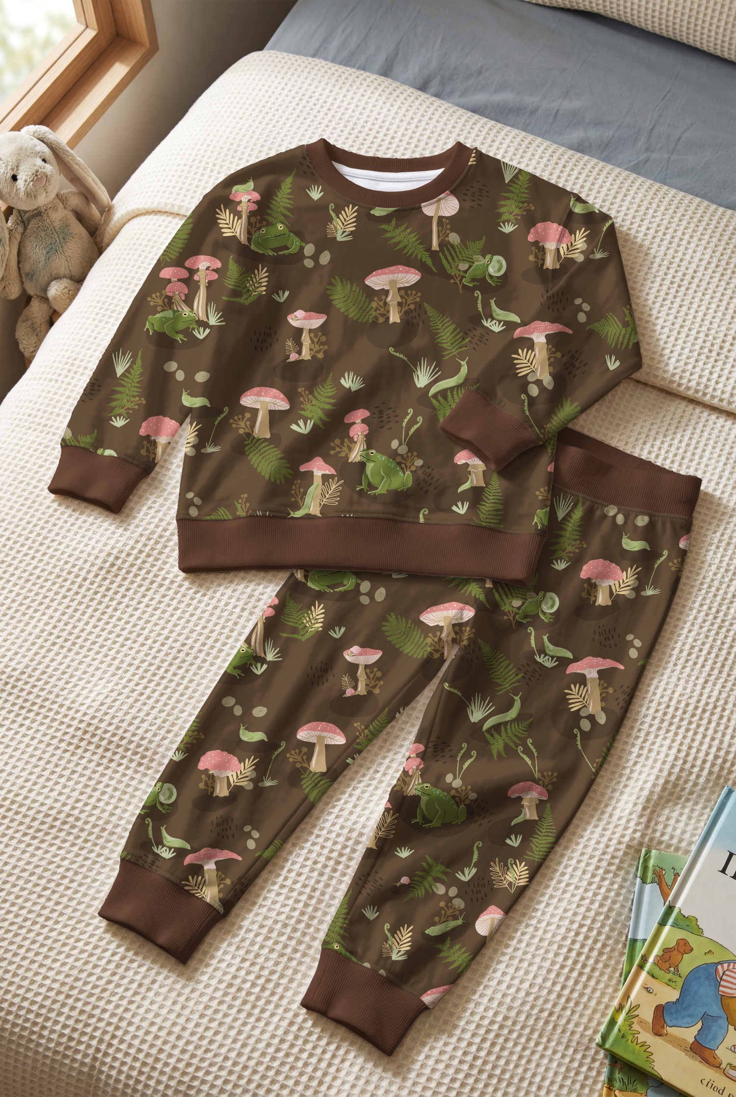 Children's pajamas with a woodland animal pattern including frogs, salamanders, mushrooms, and ferns, laid out on a bed with a blanket and a stuffed animal nearby.