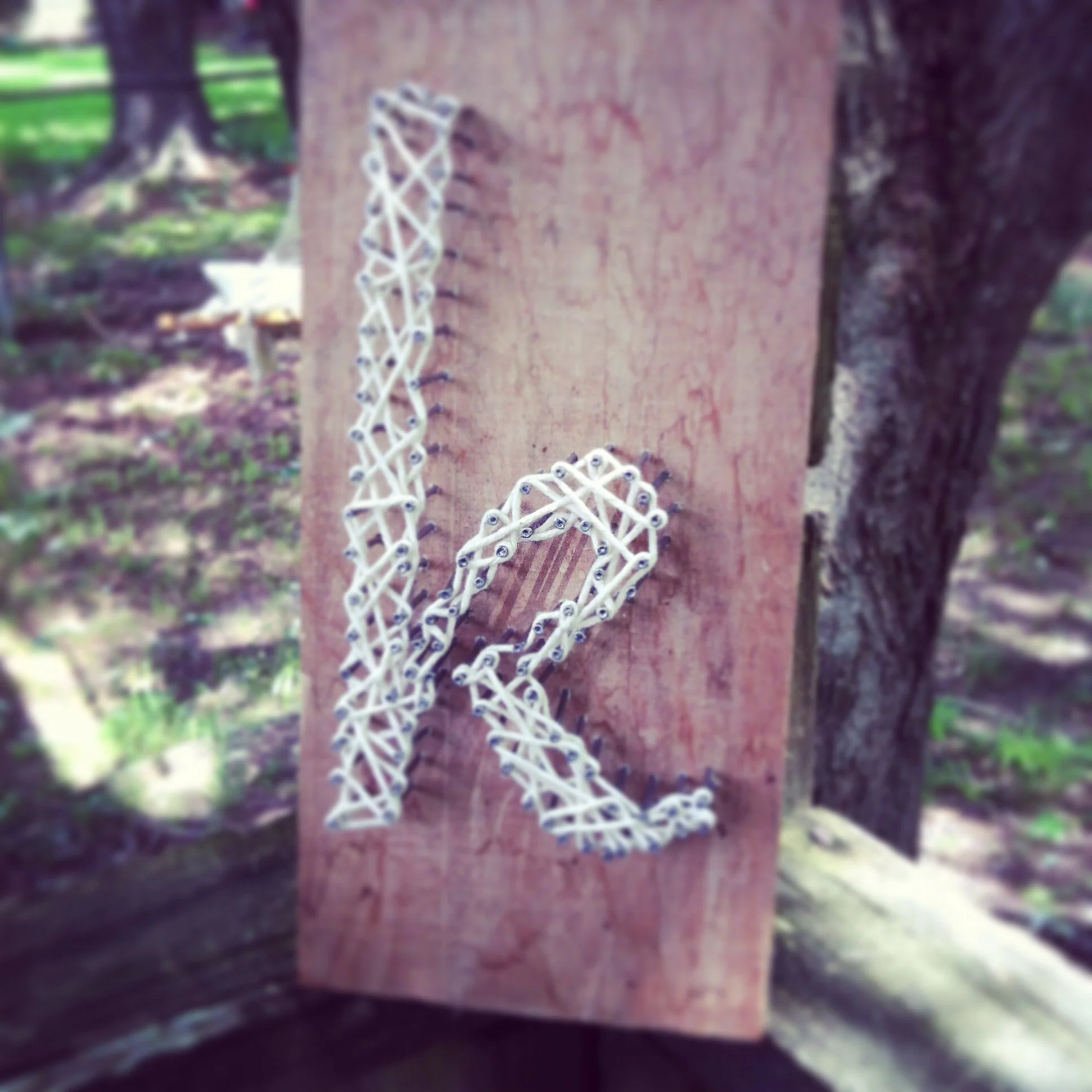 A heart shape made from white string or thread on a rough wooden surface outdoors, with blurred green foliage in the background.