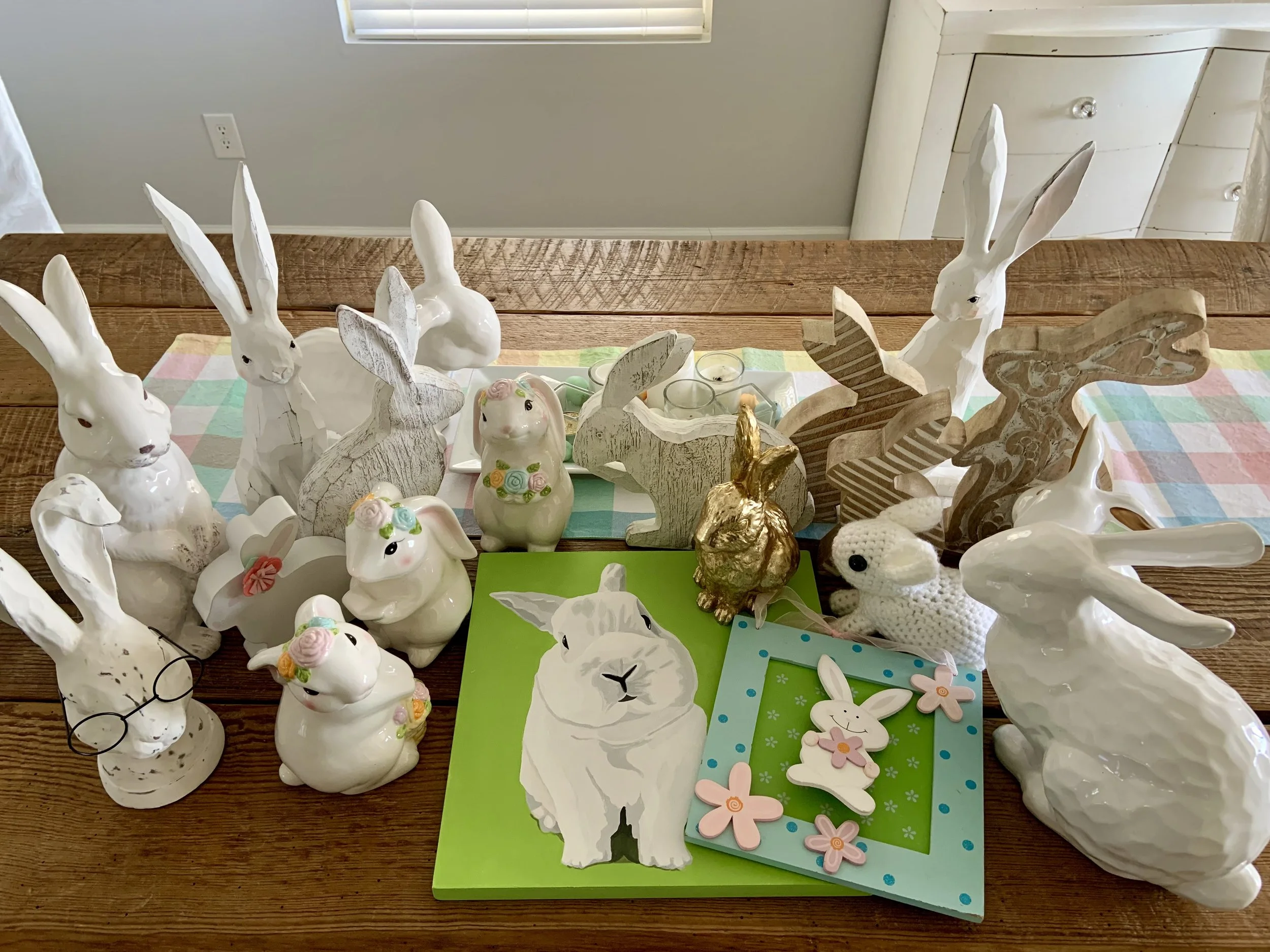 A collection of Easter-themed decorations and gifts displayed on a wooden table. The items include ceramic, wooden, and resin bunnies in white and gold, along with an original acrylic painting of a white bunny on a bright spring green background.