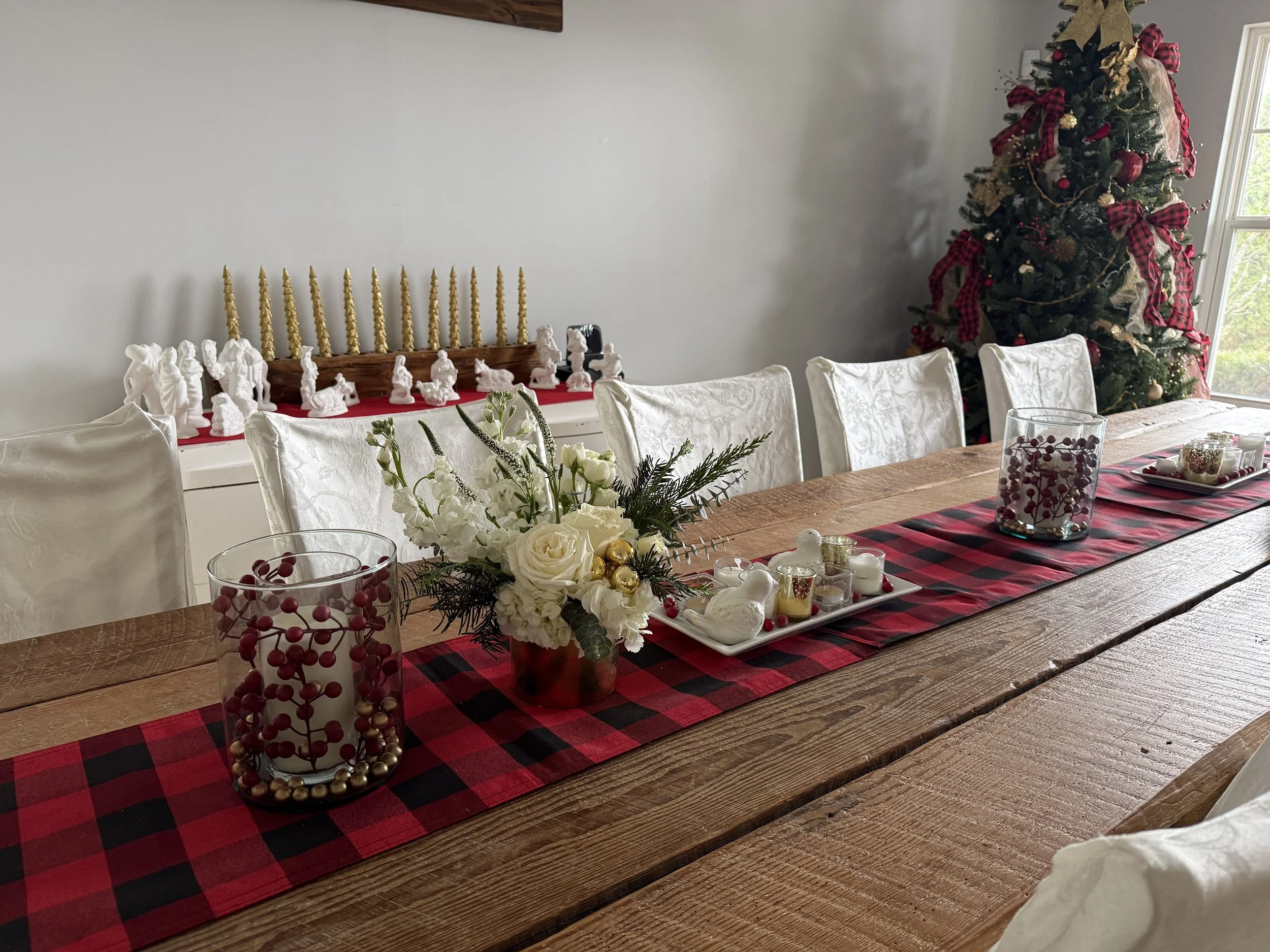 A decorated Christmas tree in a corner with red and gold ribbons, ornaments, and a gold star on top, near a window. A table with a red and black checkered table runner is set with holiday decorations, including a floral centerpiece, candles, and glass jars filled with berries and ornaments.