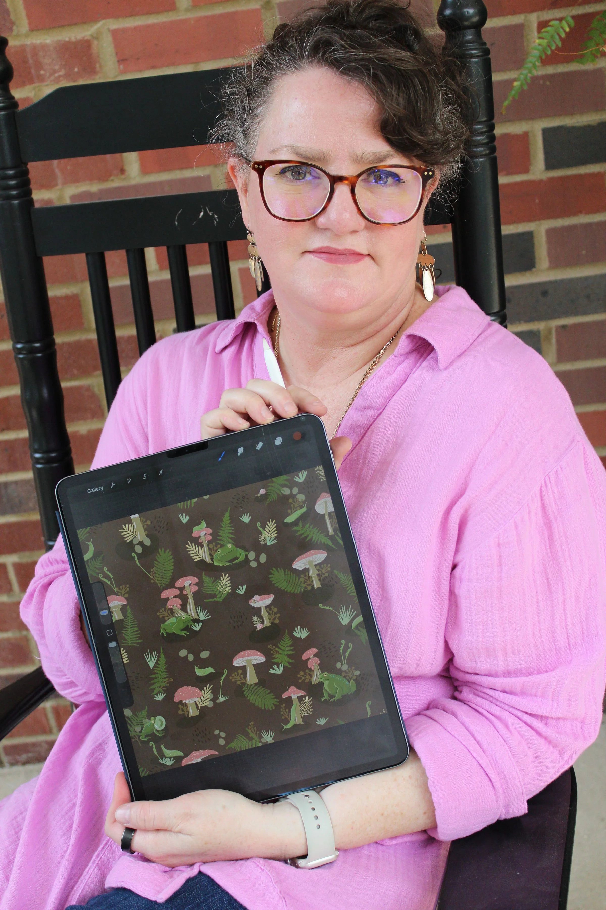 A woman with curly hair and glasses wearing a pink shirt, sitting on a black chair with a brick wall in the background, holding a tablet displaying a nature-themed illustration of various plants, mushrooms, and frogs.