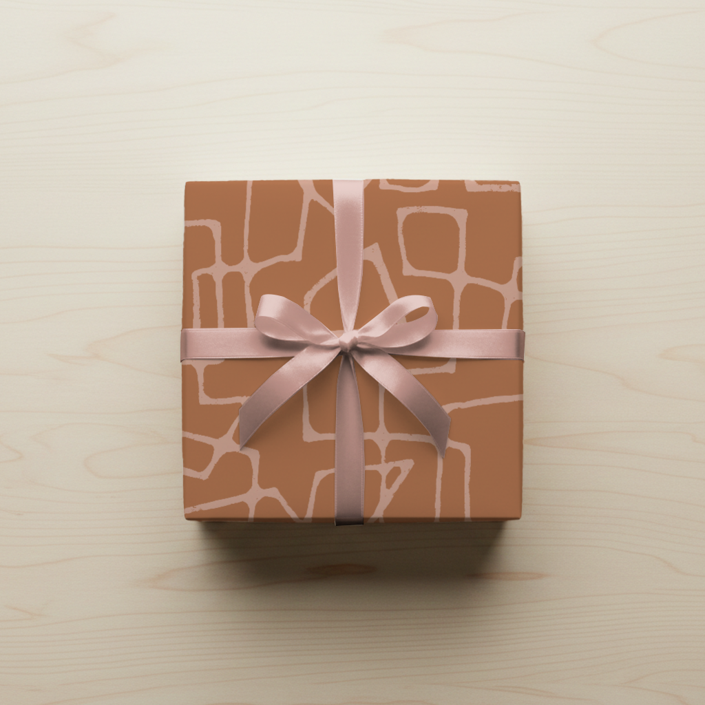 A square gift box wrapped in paper with a terracotta and pink mid-century line pattern, tied with a pink satin ribbon formed into a bow, sitting on a light wooden surface.