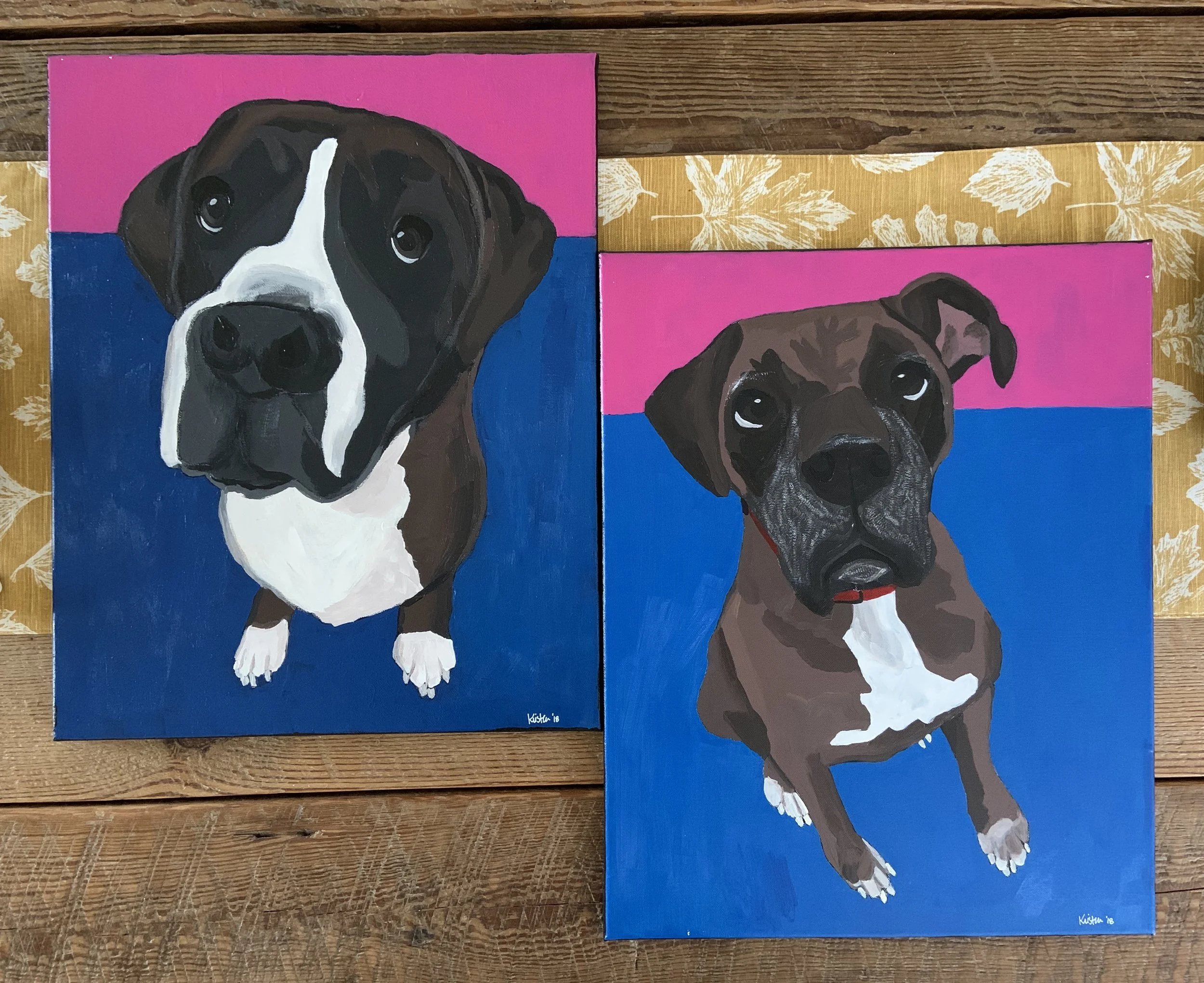 Two acrylic paintings of Boxer dogs on pink blue backgrounds, placed on a wooden table.