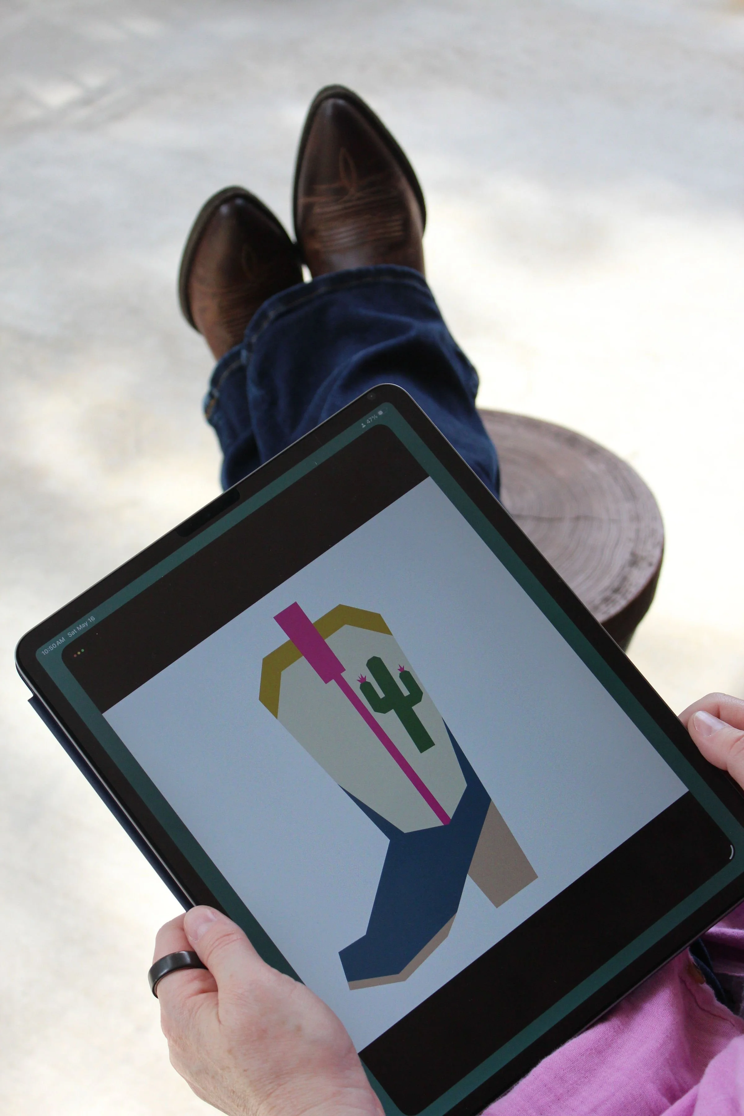 A person holding a tablet displaying a colorful graphic with a cactus. The person is wearing a pink sleeve and a black ring on their finger. In the background, there are brown cowboy boots and denim jeans, with a wooden log behind their hand.