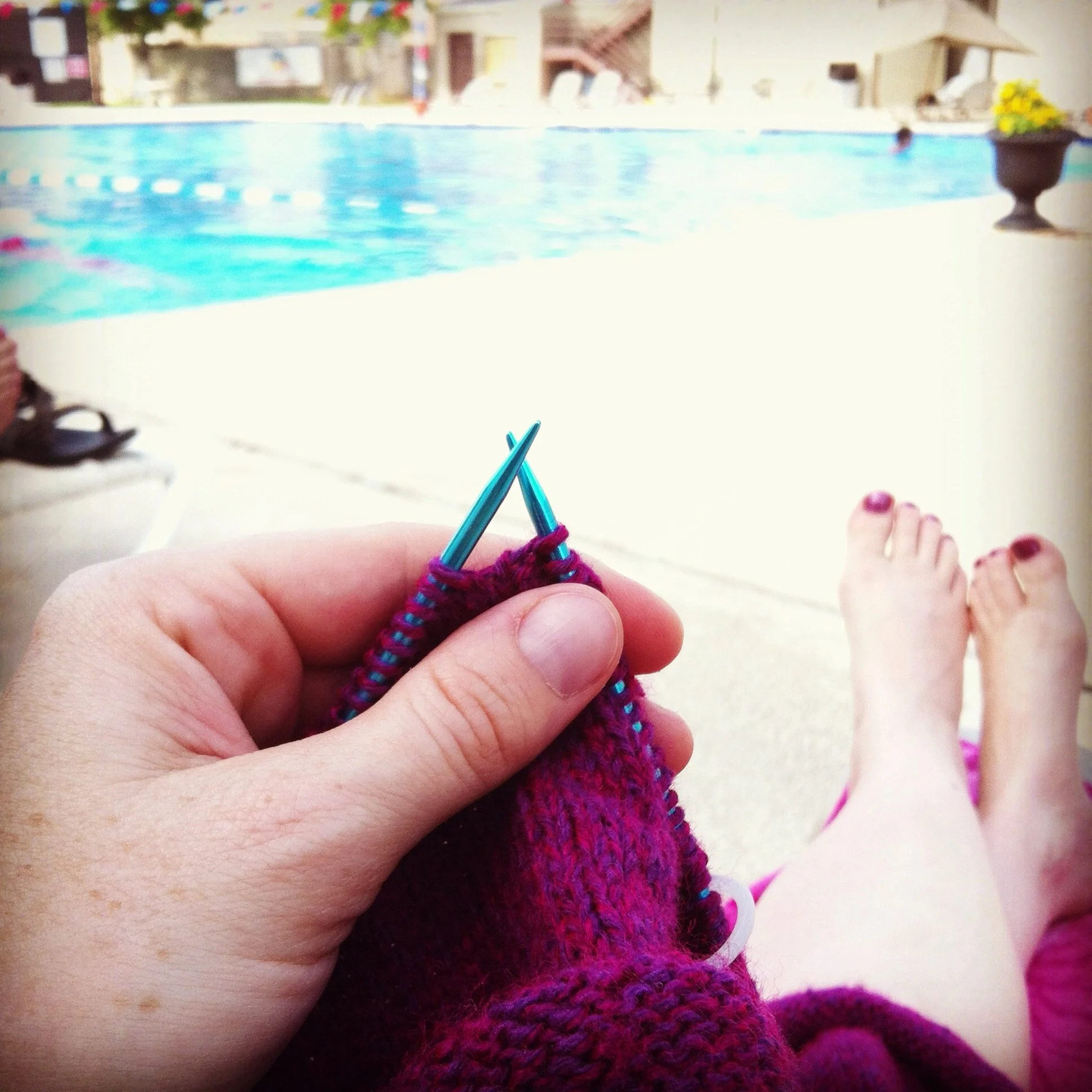 Person knitting with purple yarn on their lap by swimming pool.