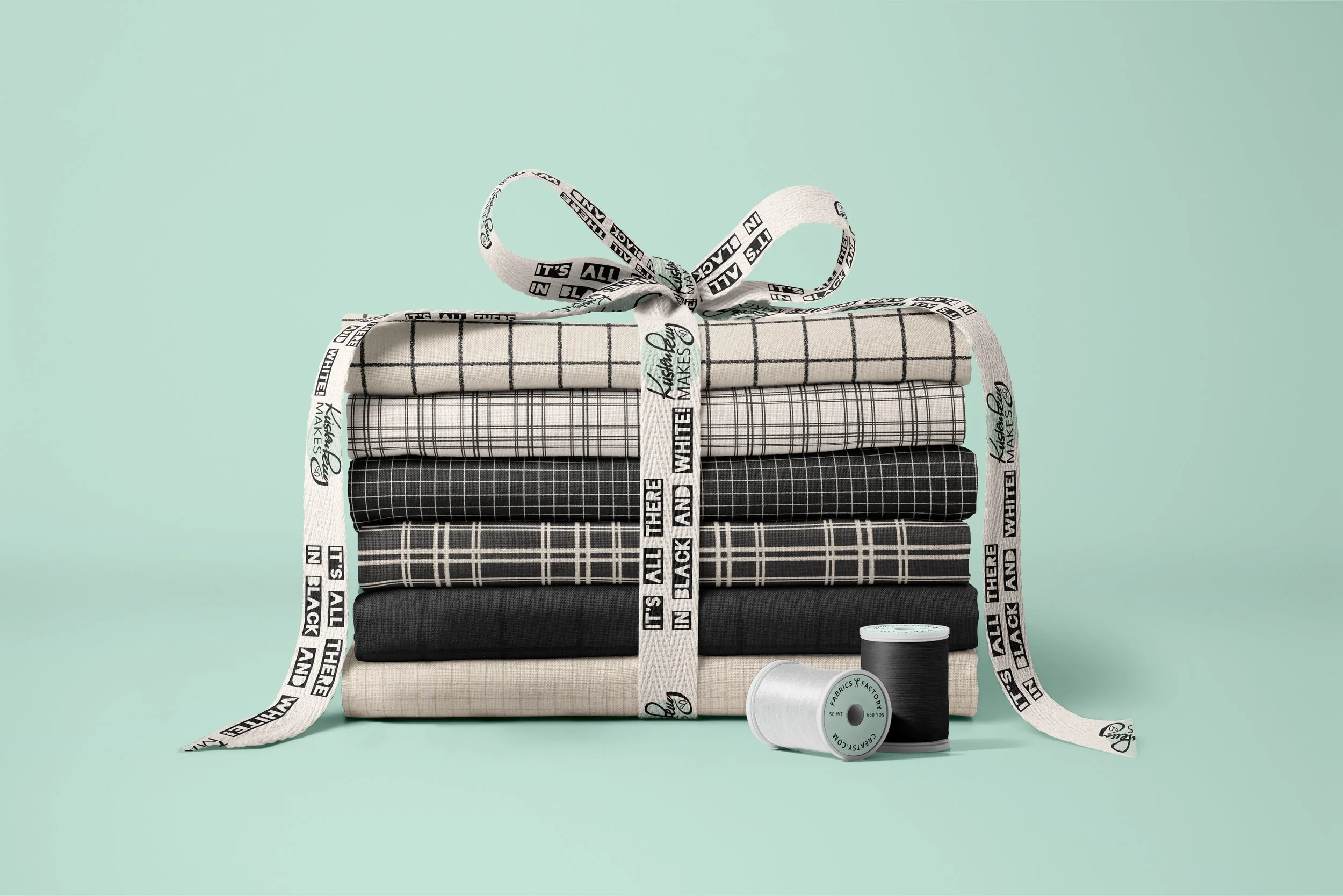 Stacked folded fabric pieces with various black, white, and gray plaid patterns, tied with a white ribbon that has black text, positioned beside black and white spools of thread on a light teal background.