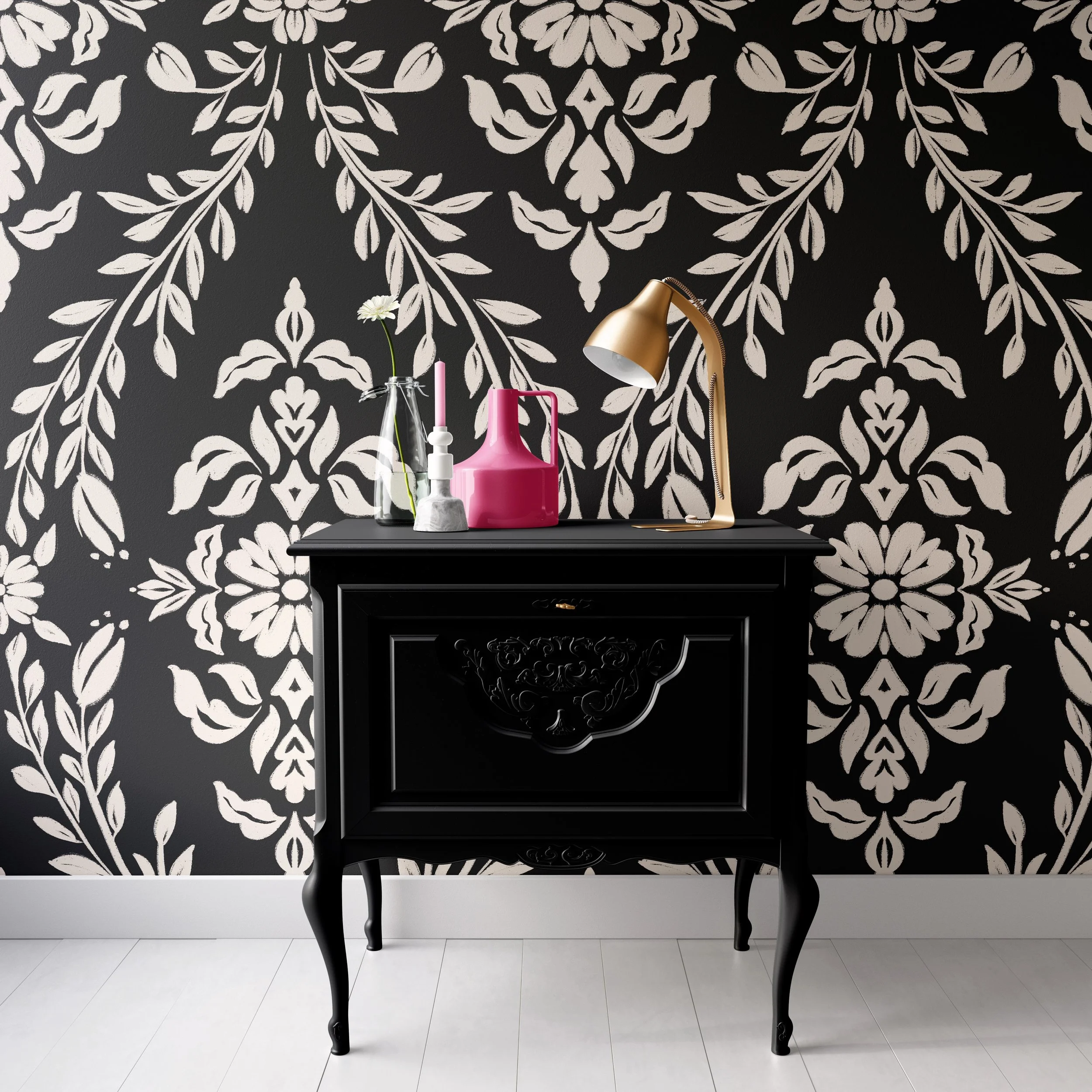 A black dresser with ornate carvings on a white floor against a black and white floral damask wallpaper background. On the dresser, there are a clear glass bottle with a white flower, a pink pitcher, and a gold desk lamp.