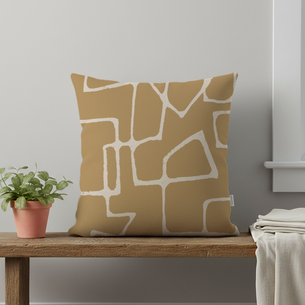 A decorative pillow with an ochre and sand abstract geometric pattern on a wooden surface, beside a potted plant with green leaves and some white folded fabric.