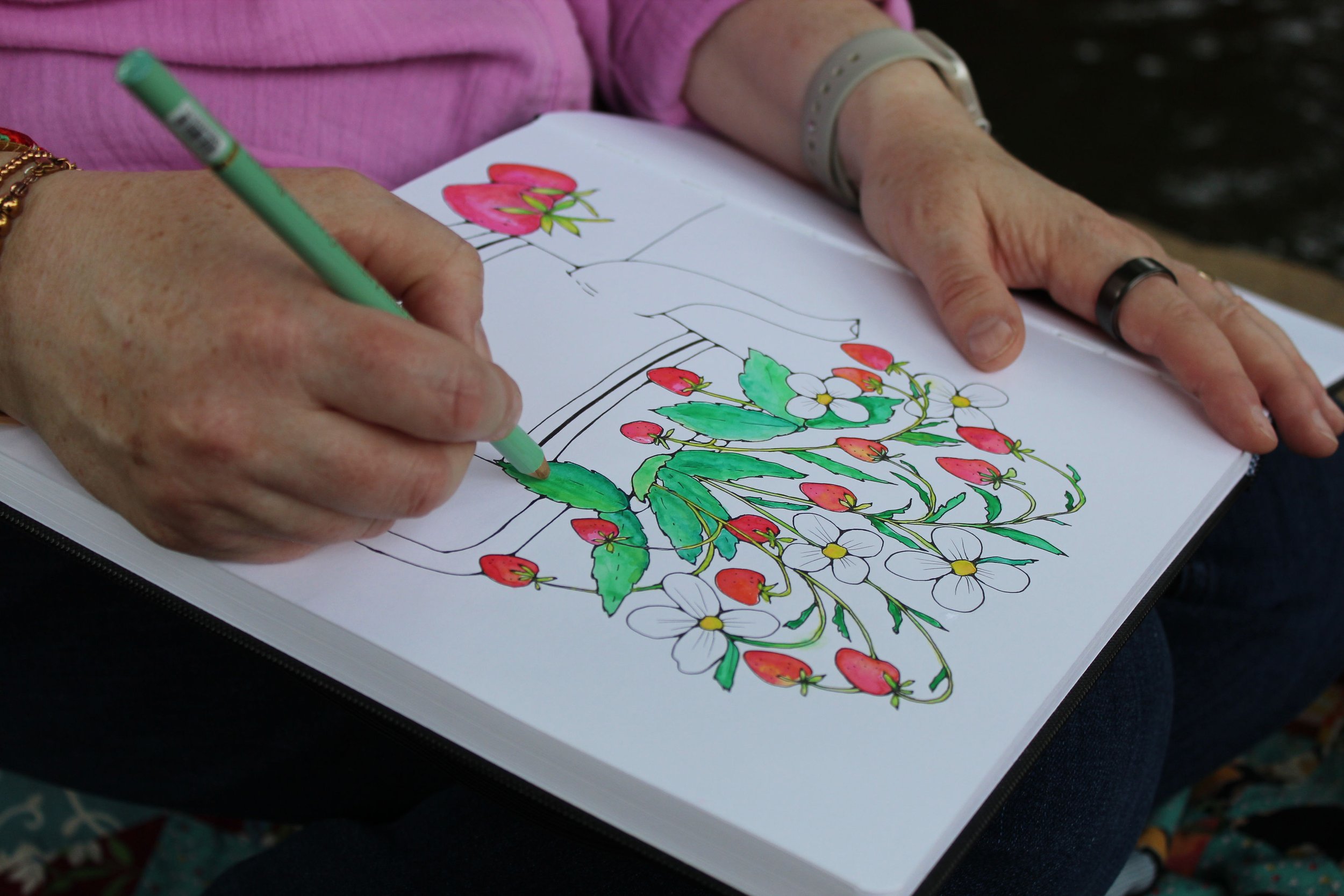 A person is coloring a botanical illustration of strawberries, leaves, and flowers in a coloring book.