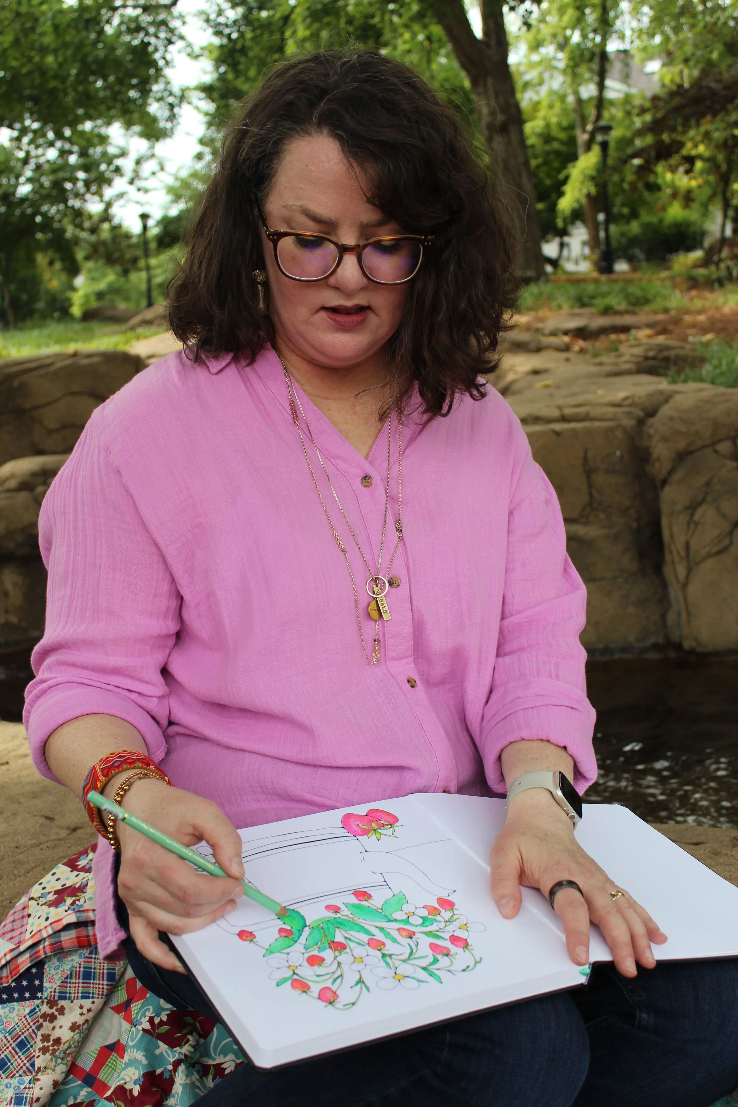 A woman with curly dark hair, wearing glasses and a pink shirt, sitting outdoors in a park with green trees in the background. She is coloring a flower drawing in a notebook.