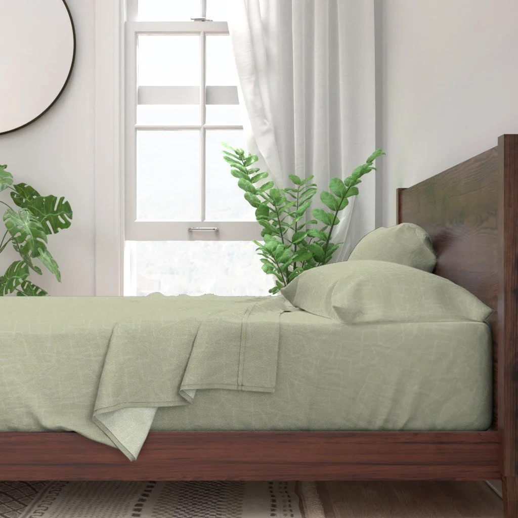 A bed with bedding and pillows featuring a pale green mid-century modern abstract tone-on-tone pattern, wooden headboard, green plants on either side, white curtains partially open, bright window in the background, and a round mirror on the wall.