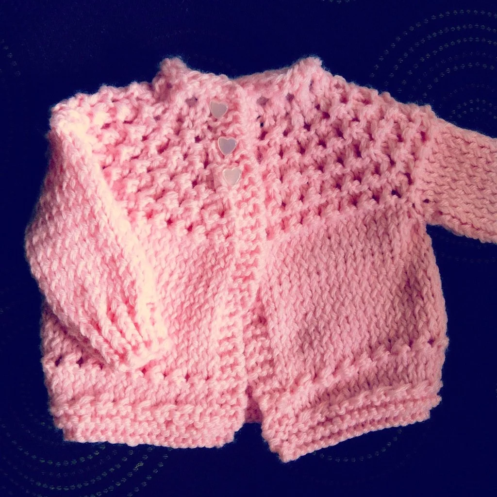 Pink knitted baby sweater with heart-shaped buttons on a black surface.