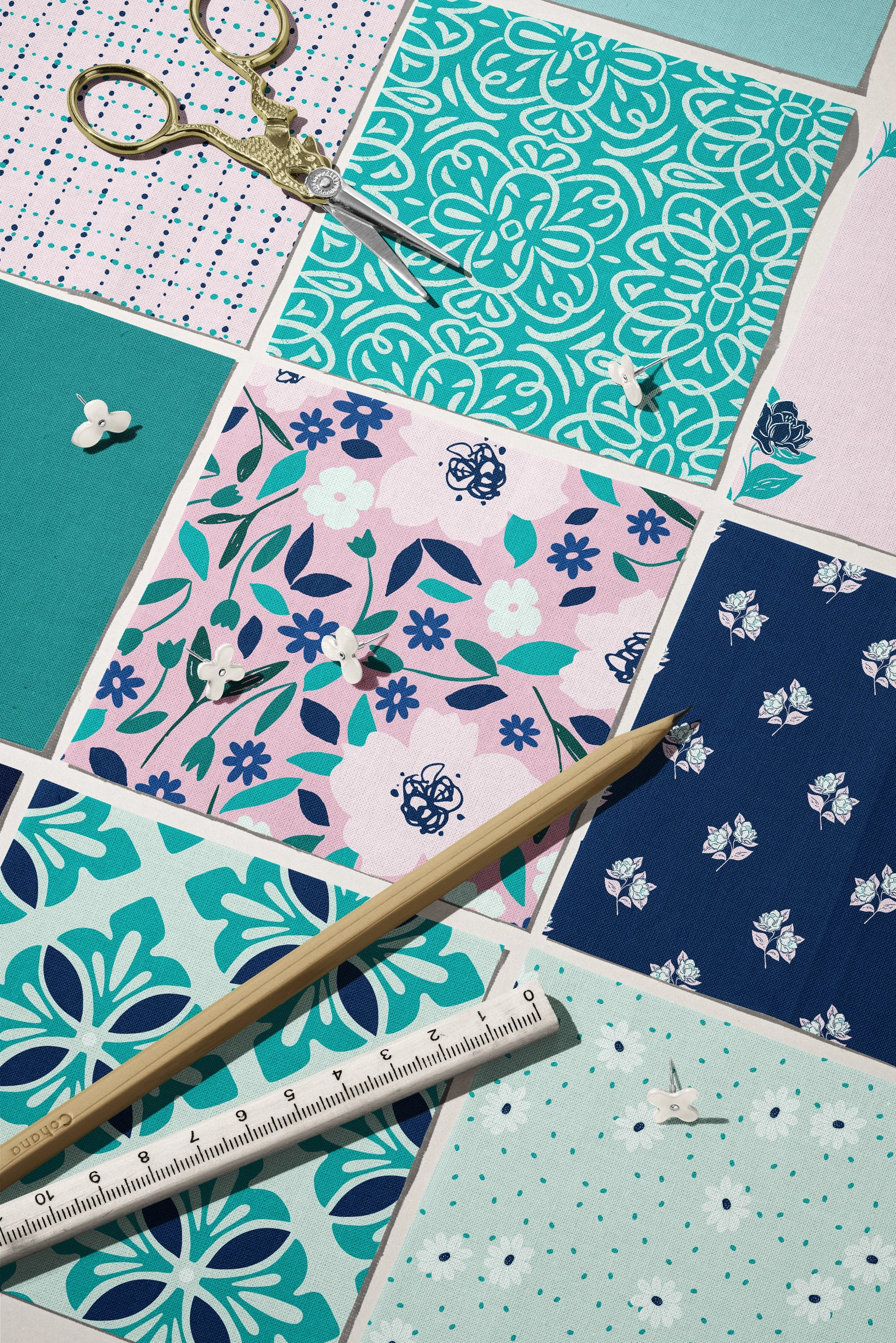 Various fabric swatches with floral and geometric patterns in shades of navy, pink, mint, and teal, along with sewing tools including a pair of gold scissors, white flower-shaped push pins, a wooden pencil, and a white ruler with black markings.