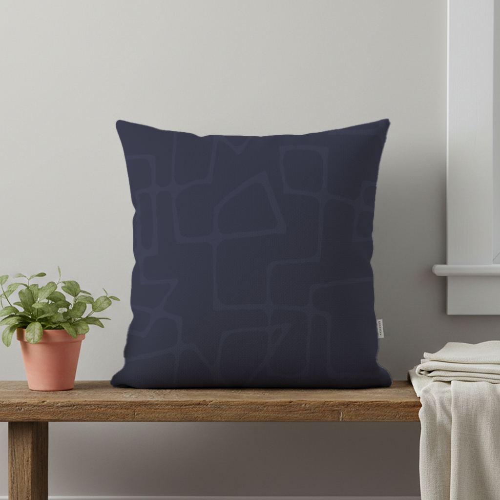 A navy blue decorative pillow with a subtle abstract mid-century modern pattern sits on a wooden surface next to a small potted plant with green leaves and a white blanket.