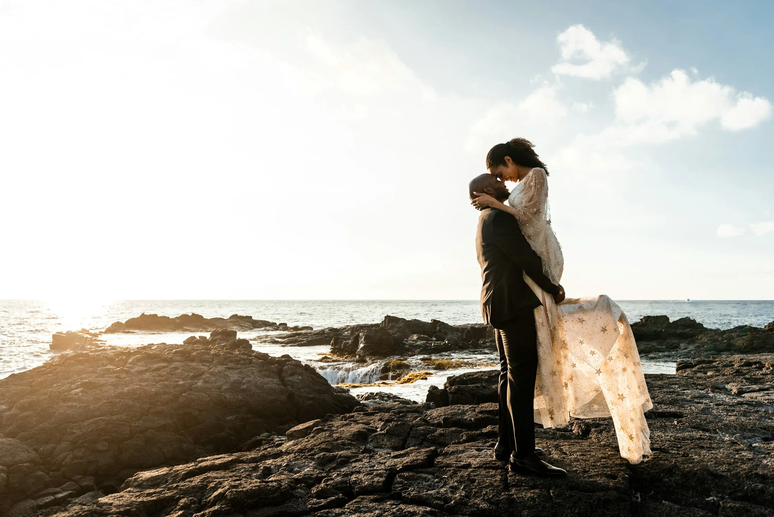 Romantic elopement couple embracing on rocky ocean shore at sunset