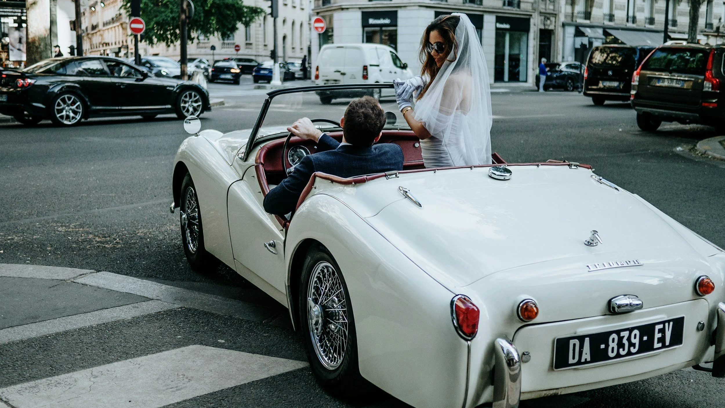 Vintage wedding scene with bride and groom in classic Triumph convertible, highlighting retro elegance and charm.