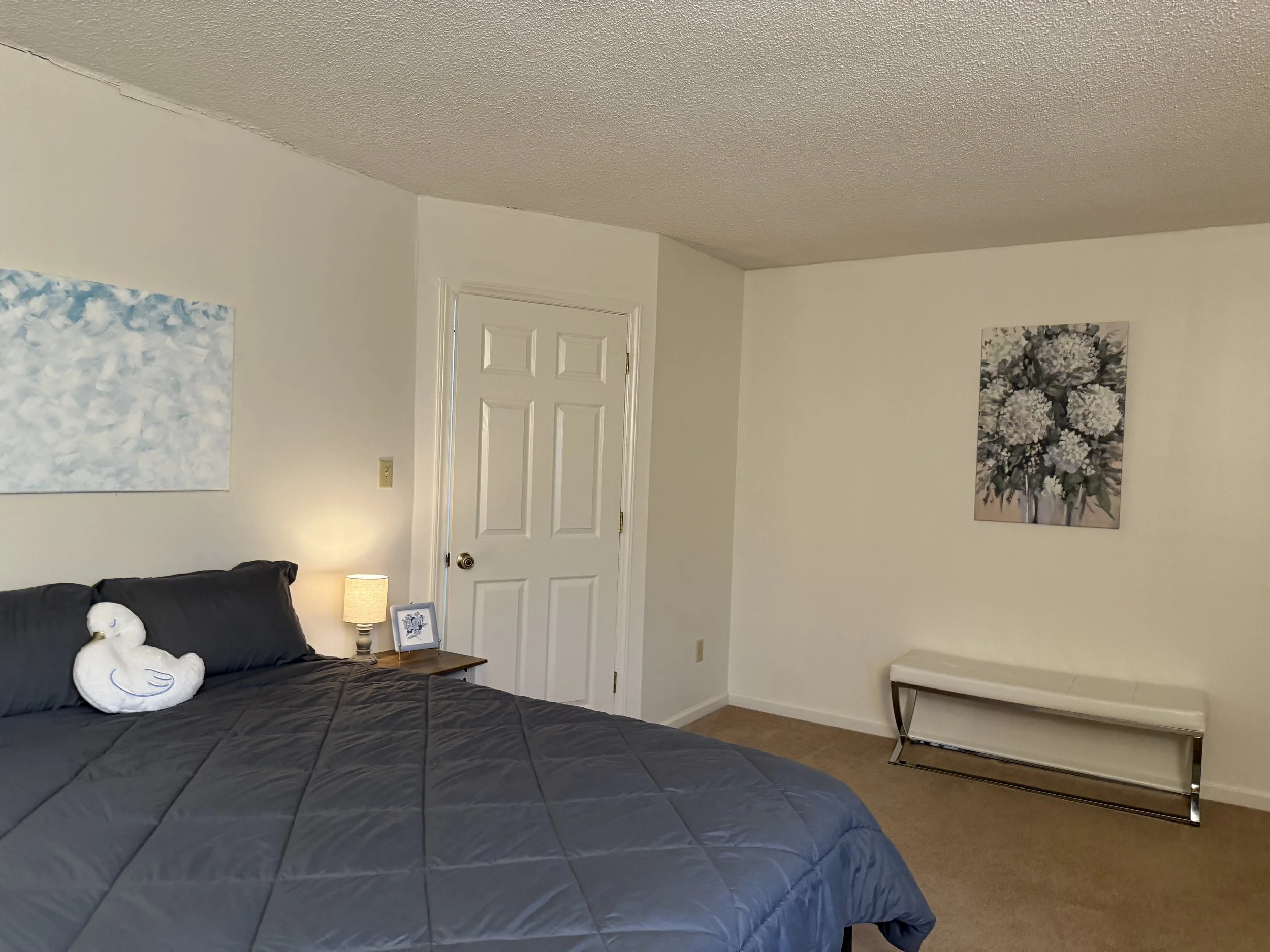 A bedroom with a bed covered in a black quilt, a white fluffy swan pillow, a bedside table with a lamp, framed artwork of a flower bouquet, a closed white door, and a white bench at the foot of the bed.