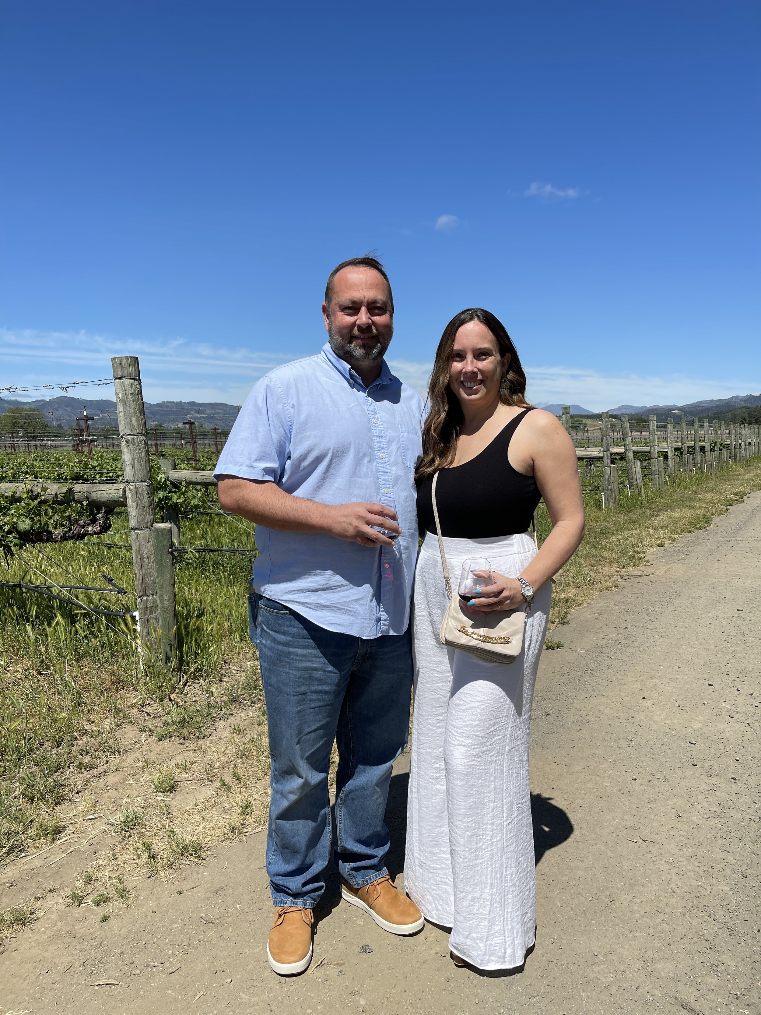 A man and a woman standing outdoors on a sunny day, holding drinks and smiling. They are in a rural area with a vineyard in the background.
