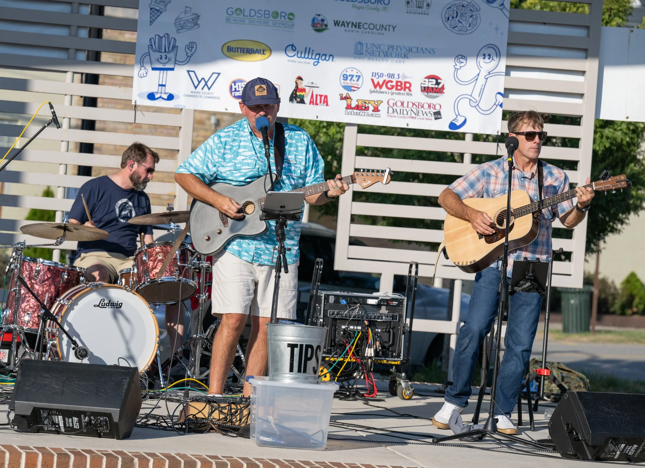 A three-member band performing live on an outdoor stage with umbrellas and a banner sponsored by local businesses in the background. The band includes a drummer, a guitarist singing into a microphone, and a second guitarist singing and playing acoustic guitar. The setting suggests a community event or festival.
