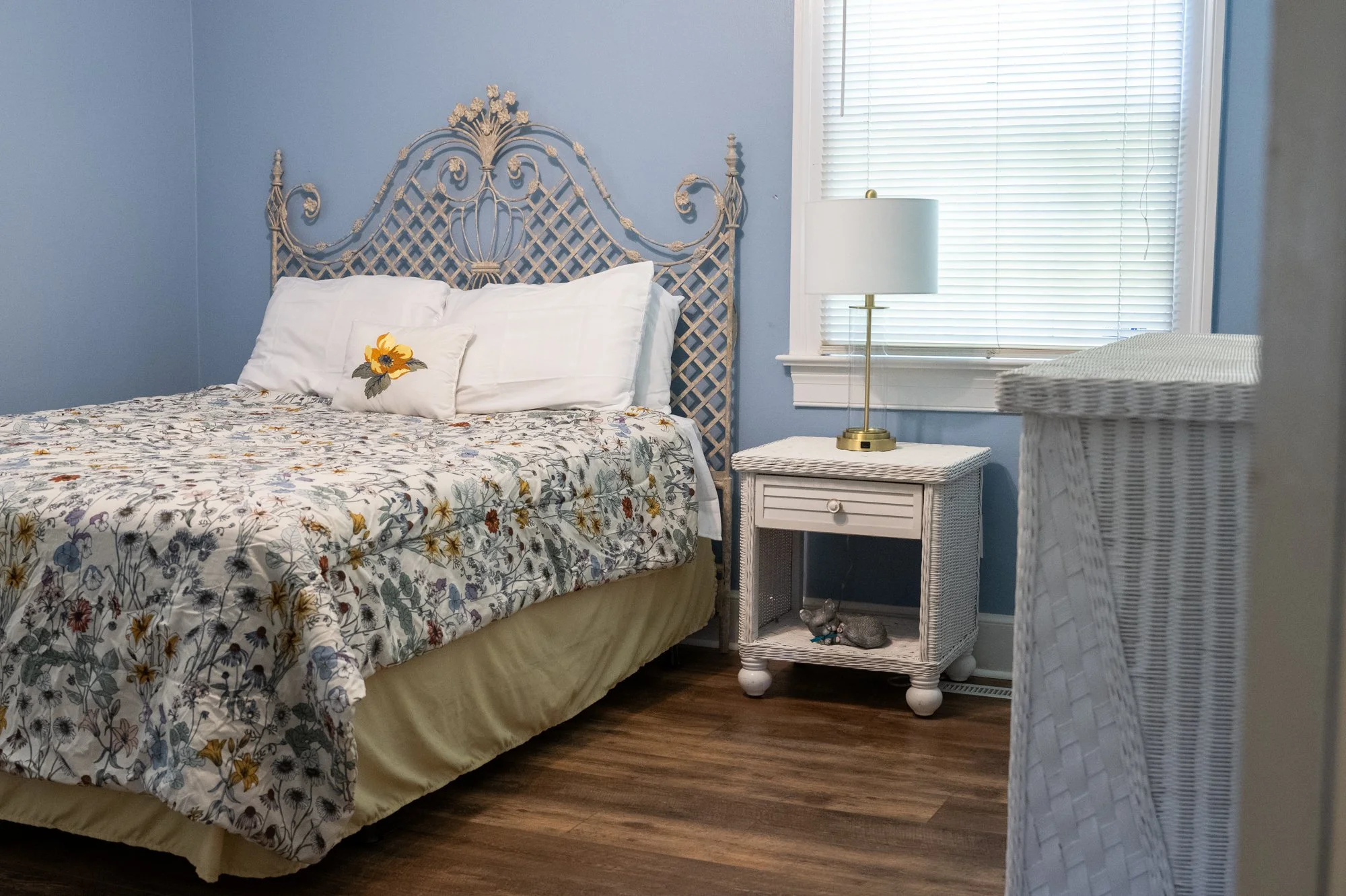 A bedroom with a white wicker headboard, floral bedspread, white pillows, a white wicker nightstand with a brass lamp, a window with blinds, and a white wicker dresser.