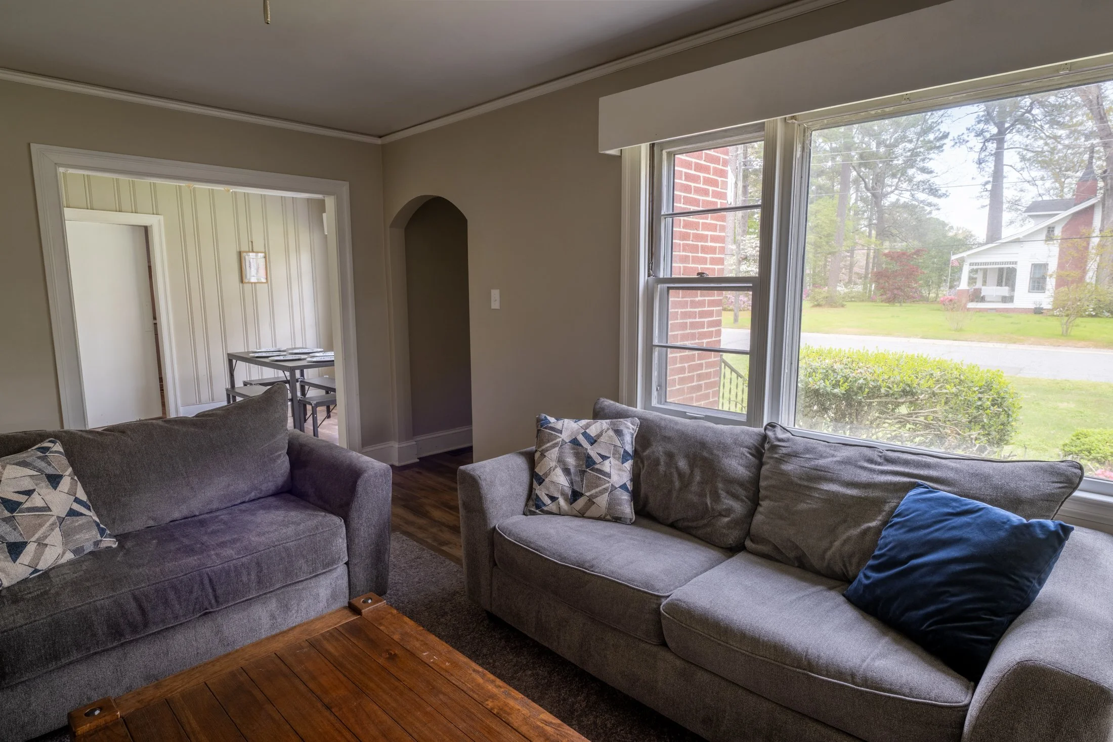 Living room with two gray sofas and patterned throw pillows, large windows overlooking a suburban yard, opening to a dining area with a table and chairs.