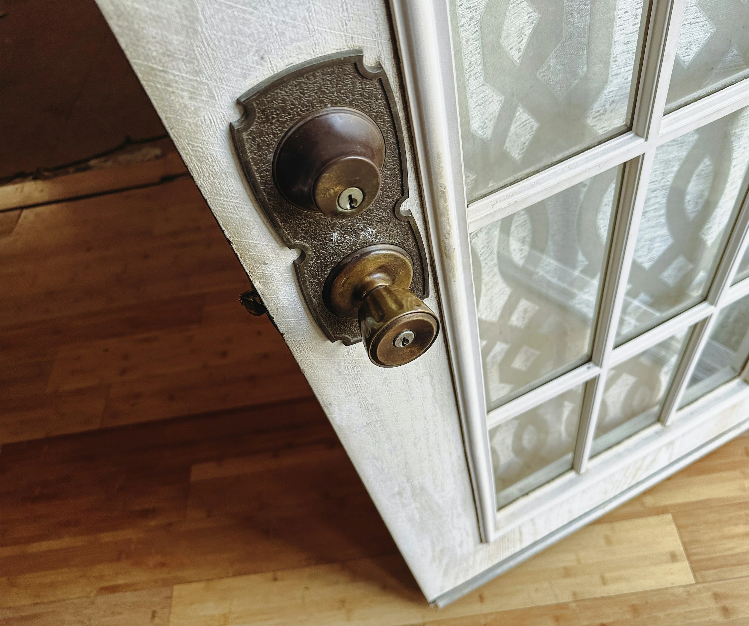 Close-up of a vintage brass doorknob with a lock on a white wooden door with glass panels, wooden floor visible.
