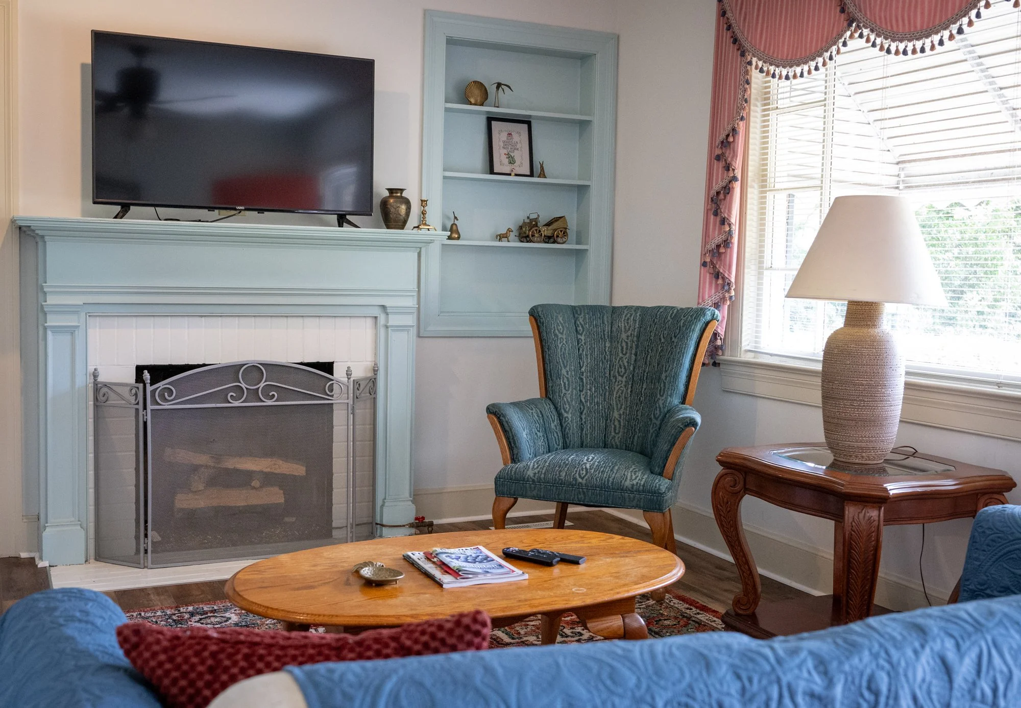 Living room with a teal armchair, a wooden side table with a lamp, a wooden coffee table with magazines and remote controls, a fireplace with a light blue mantel, a wall-mounted flat-screen TV, a built-in bookshelf, and a window with pink curtains.