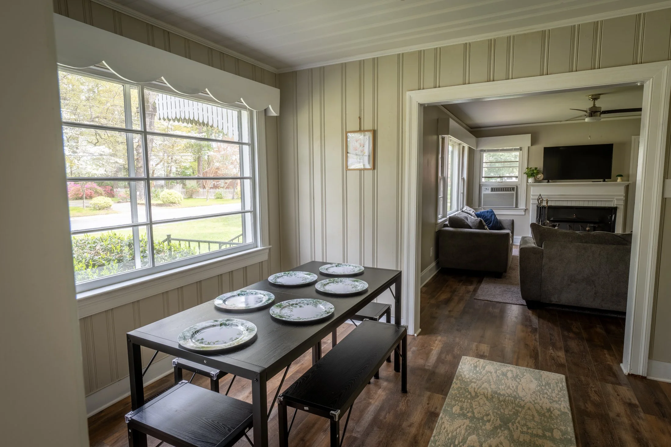 Dining area with a table set with six decorative plates near a large window, overlooking a garden. The neighboring living room features gray sofas, a fireplace, and a wall-mounted TV.