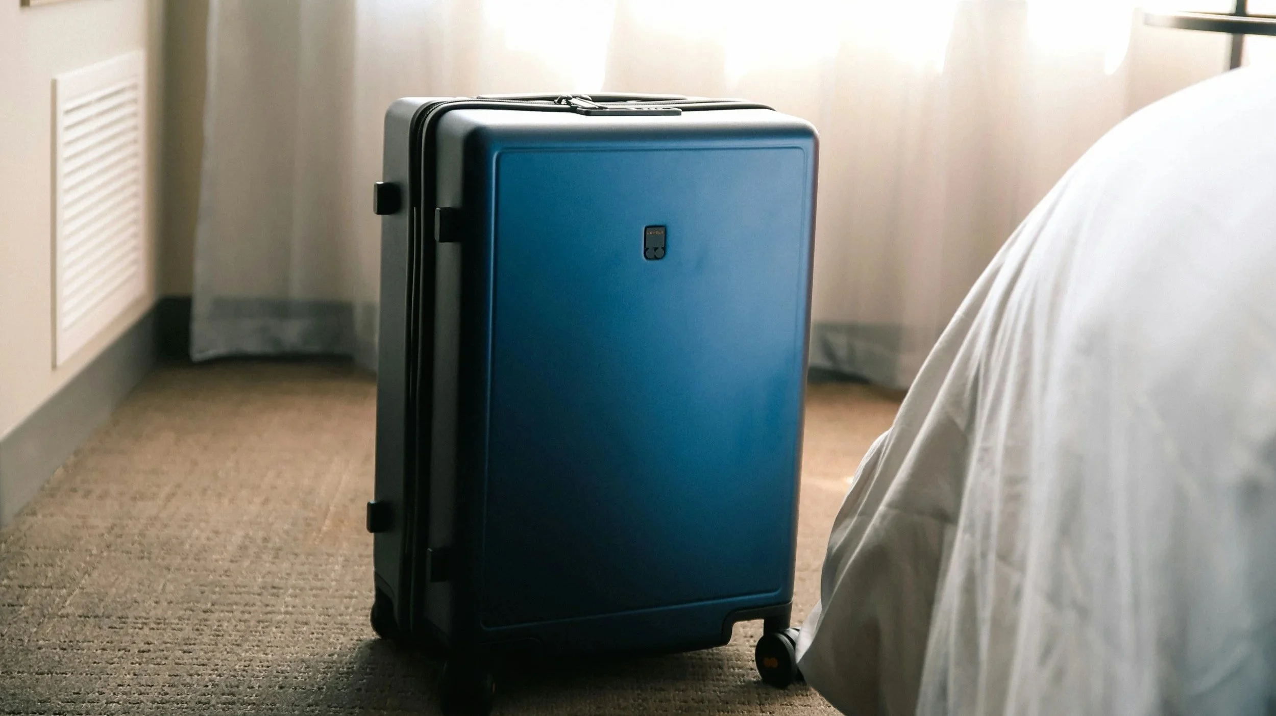 A black and blue suitcase on wheels next to a bed in a hotel room.