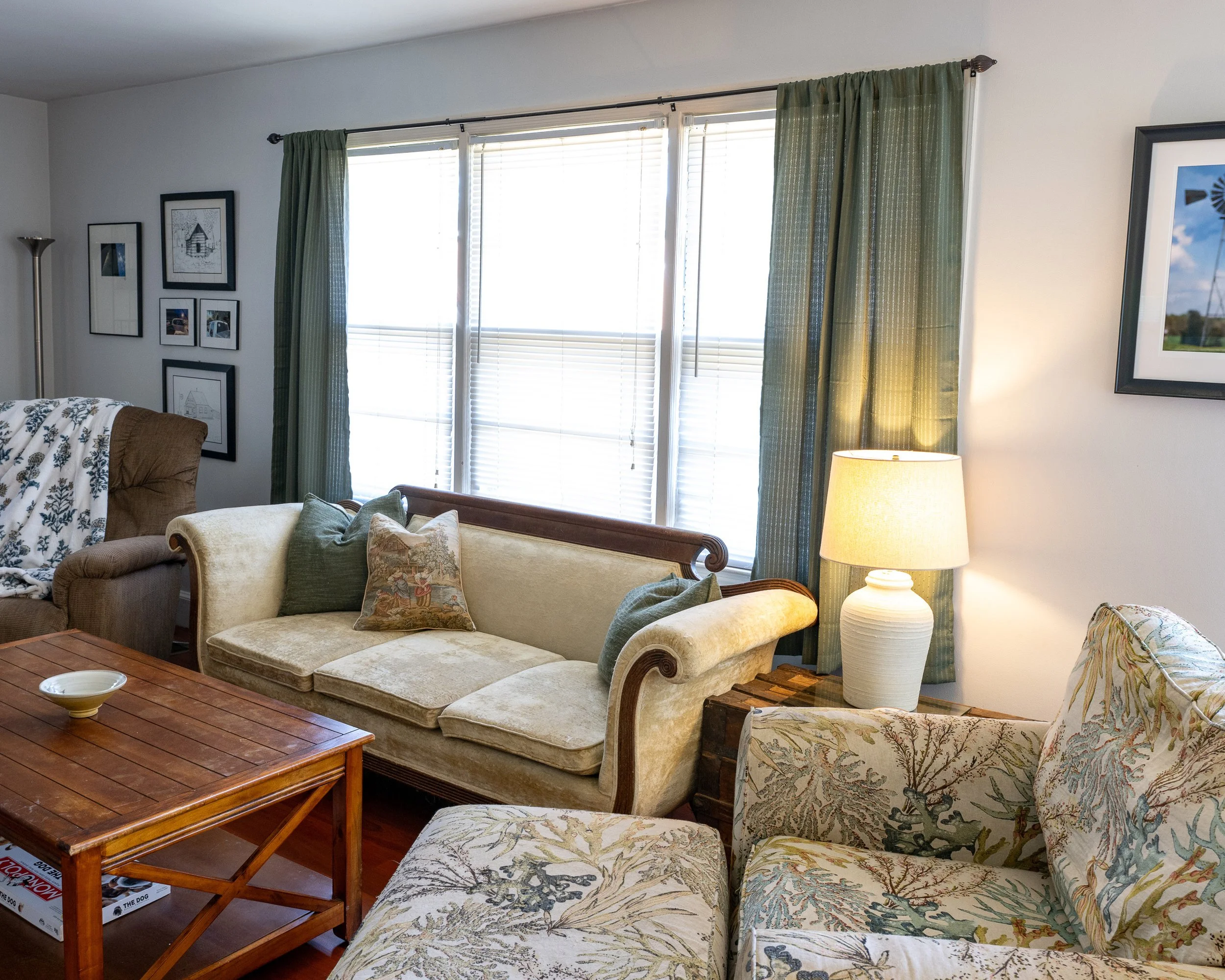 Living room with a beige sofa, patterned armchair, wooden coffee table, framed pictures on the wall, green curtains, and a table lamp.