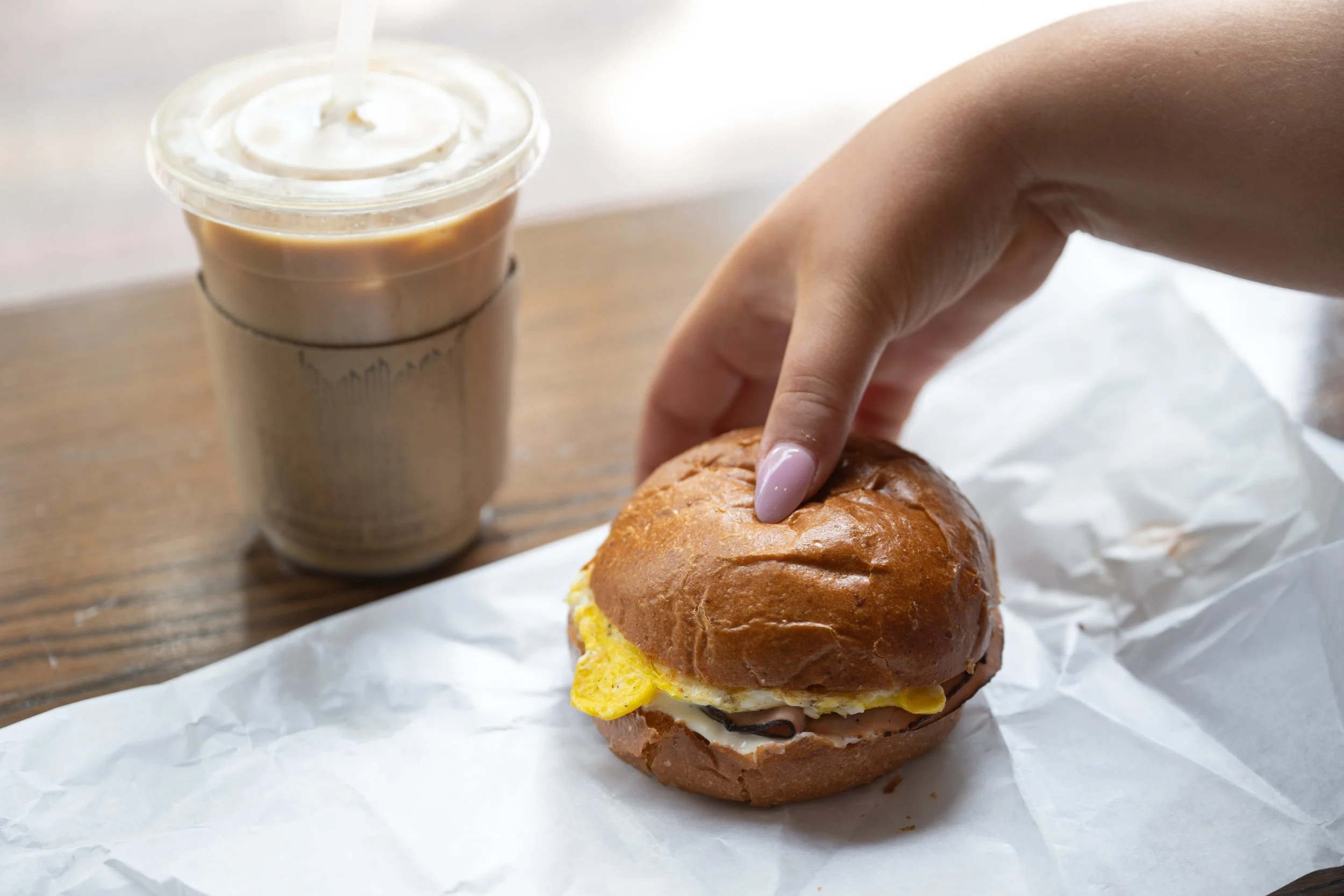 A hand holding a breakfast sandwich with scrambled eggs, cheese, and ham in a soft bun, next to an iced coffee in a paper sleeve.