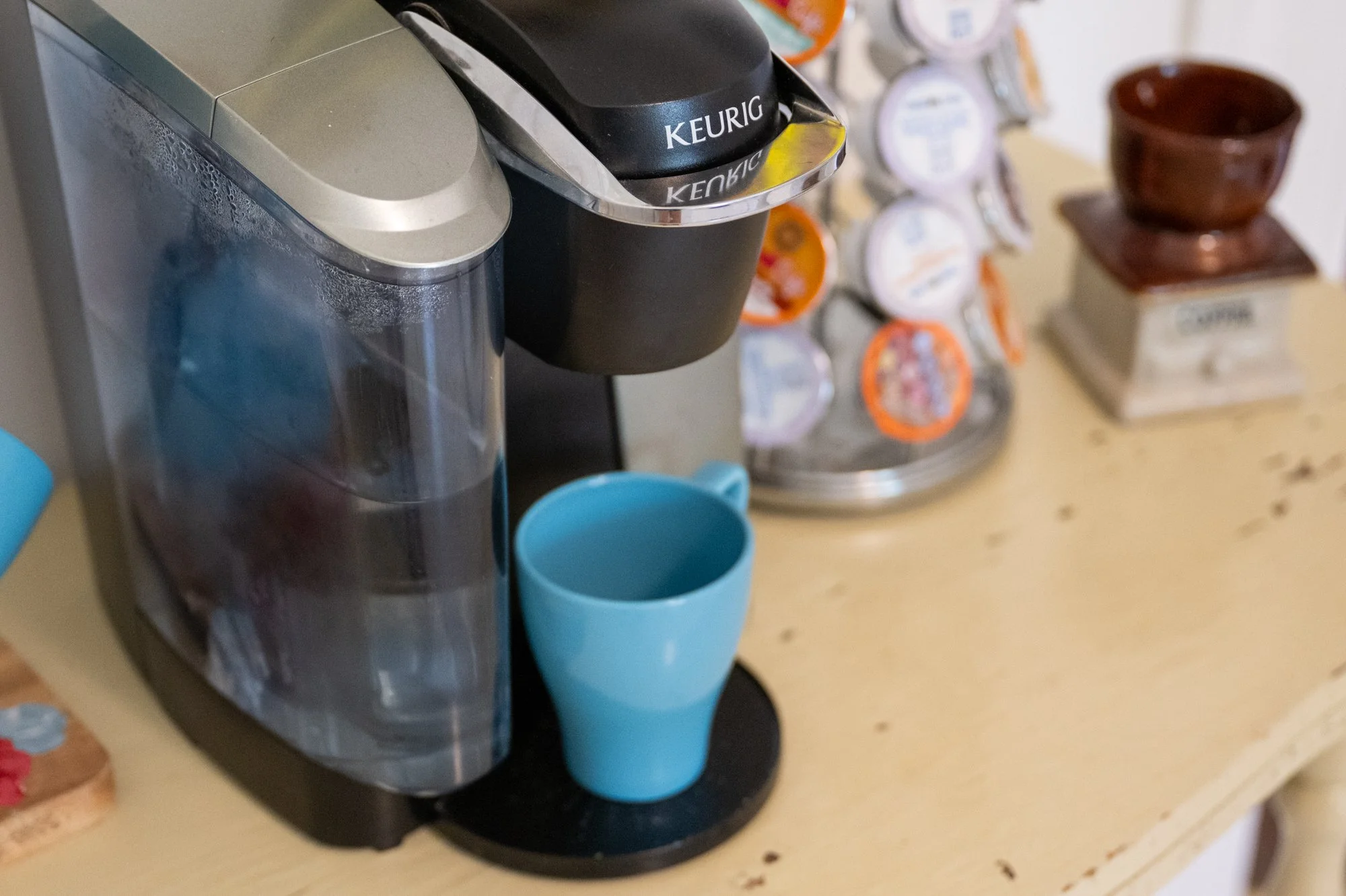 A Keurig coffee maker brewing coffee into a blue mug on a kitchen counter, with assorted K-Cup pods and dishes in the background.