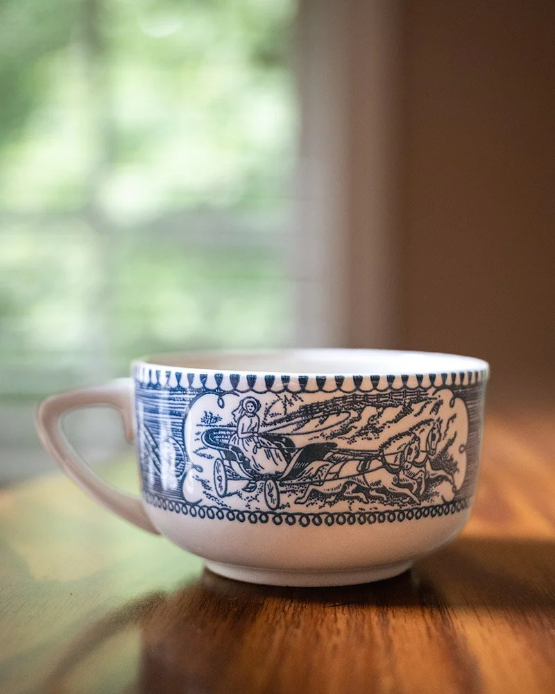 A ceramic cup with a blue and white historical or vintage illustration of a chariot drawn by horses, with a person riding inside.