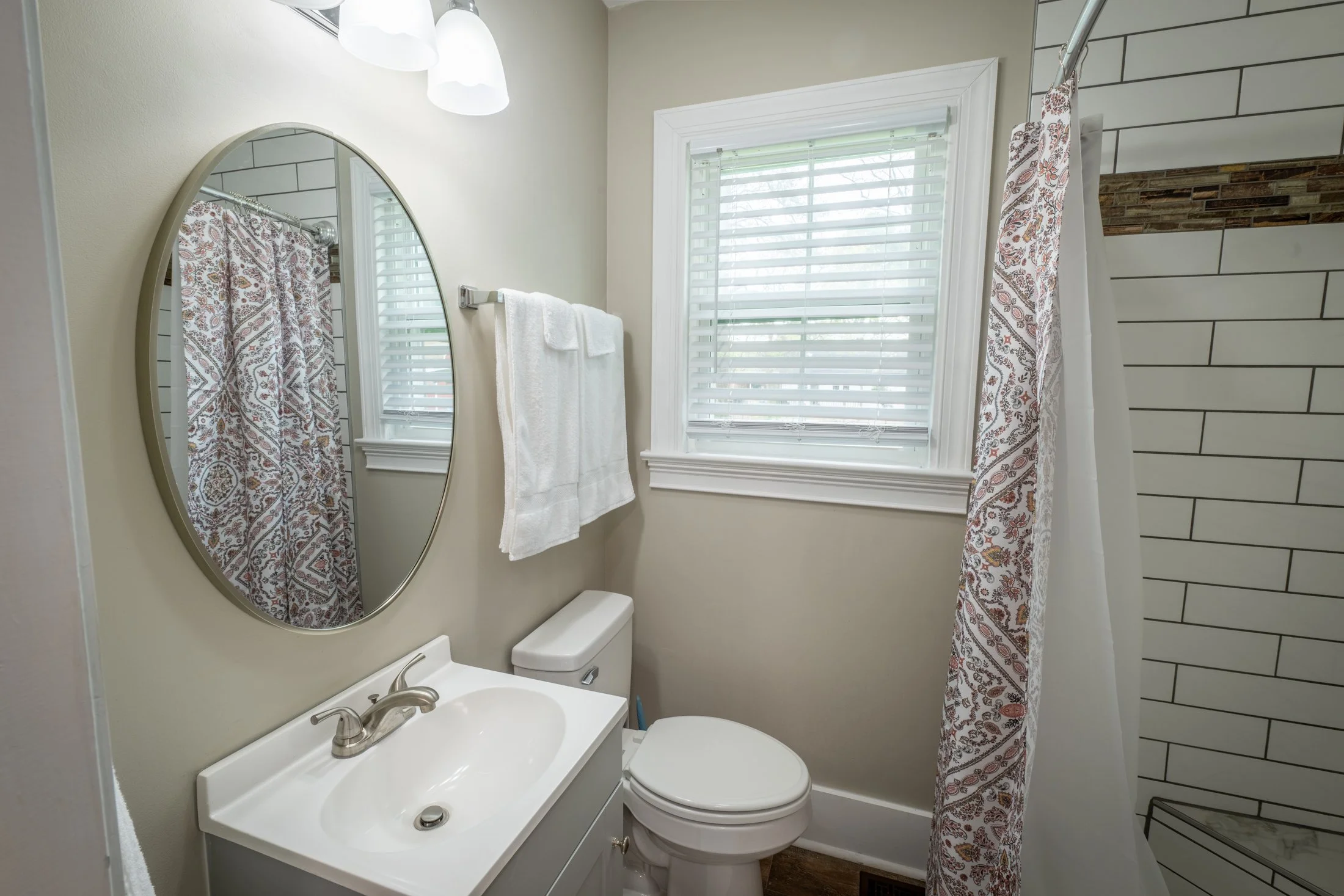 A small bathroom with a white sink, a mirror, a toilet, and a window with white blinds. There are white towels hanging on a rack, and a patterned shower curtain partially visible next to a tiled shower wall.