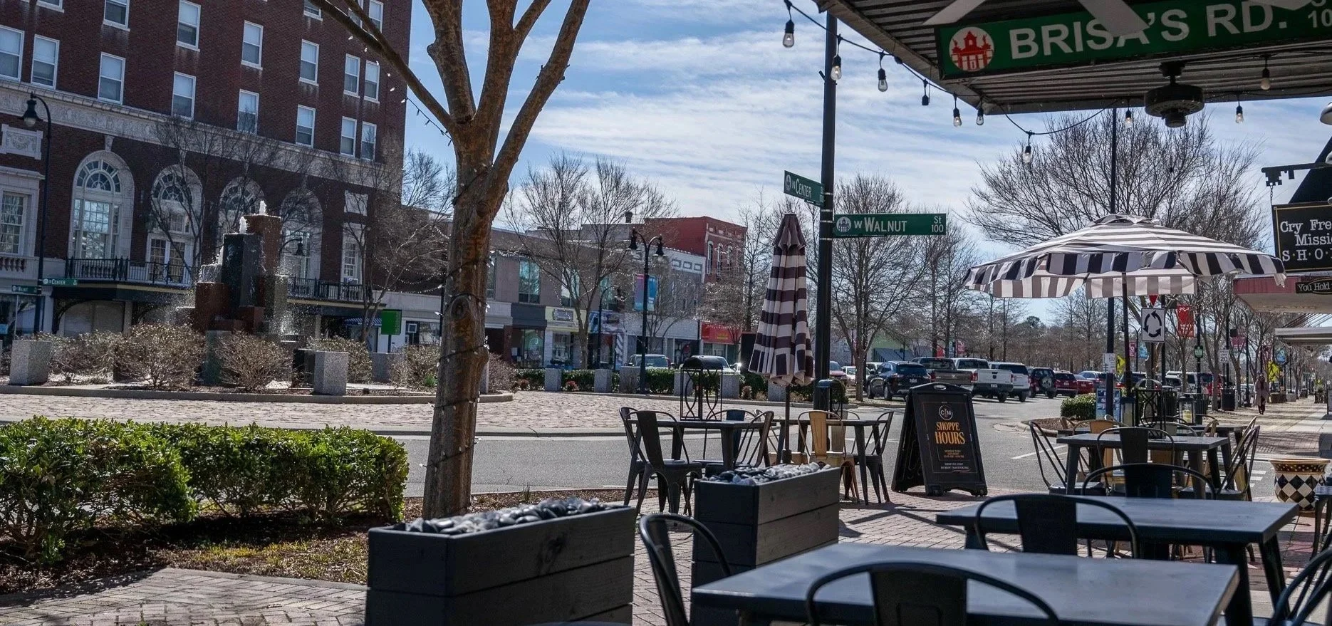 Outdoor cafe on a city sidewalk with tables, chairs, umbrellas, trees, street signs, parked cars, and buildings under a partly cloudy sky.