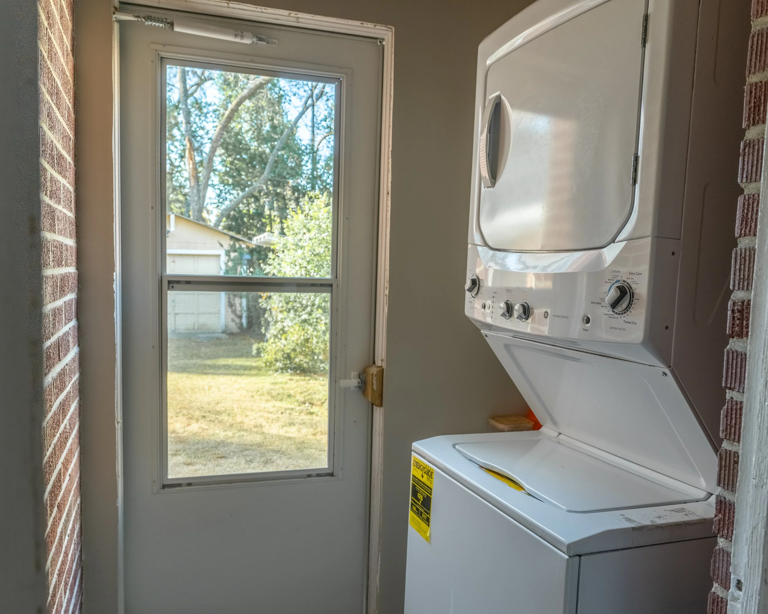 A laundry area with a white stacked washer and dryer next to a door with a window. outside view shows trees and a house in the yard.