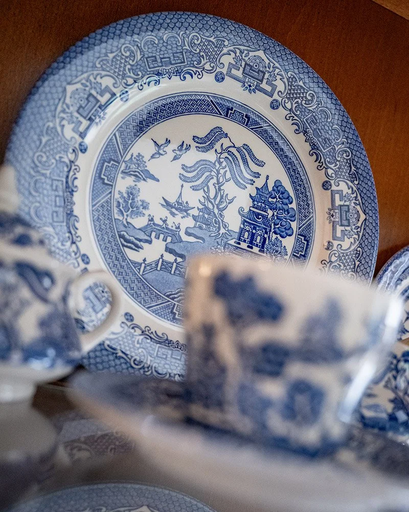 A decorative blue and white porcelain plate with a landscape scene, including trees, a pagoda, and flying birds, with teacups and saucers in the foreground.